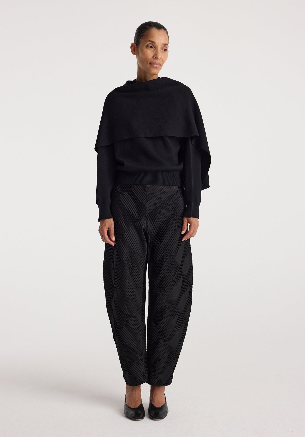 Woman wearing a stylish black cape jumper | black paired with textured black pants and black shoes, standing against a plain white background.
