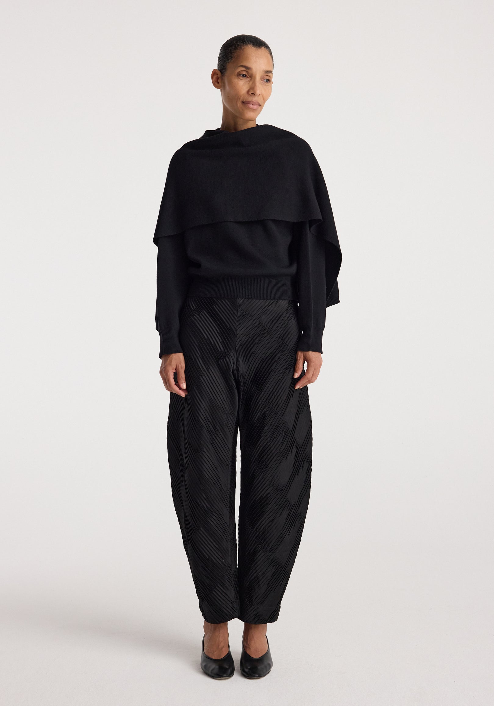 Woman wearing a stylish black cape jumper | black paired with textured black pants and black shoes, standing against a plain white background.