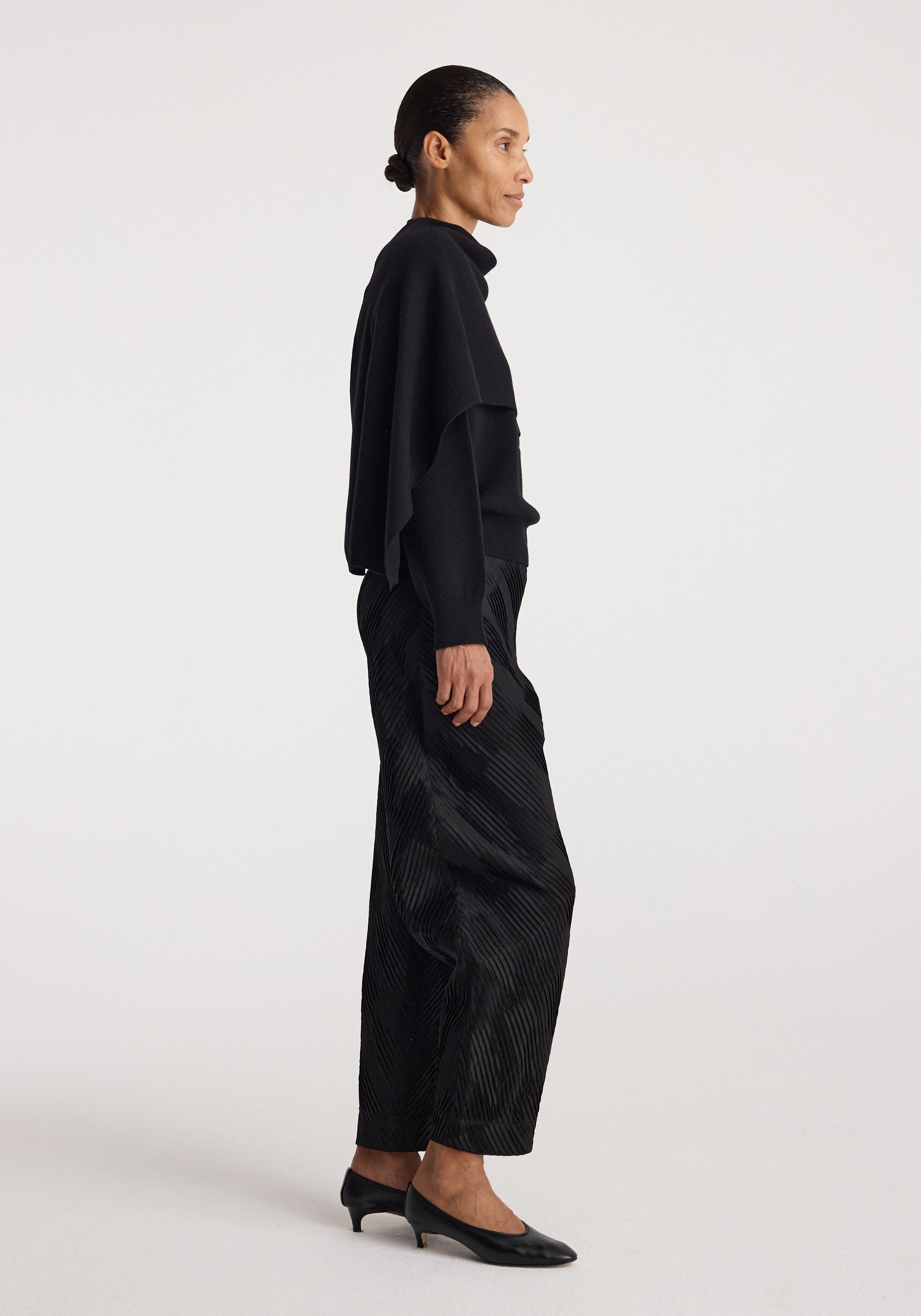 Side profile of a woman wearing the black Cape jumper | black paired with black wide leg pants and low-heeled black shoes against a plain background.