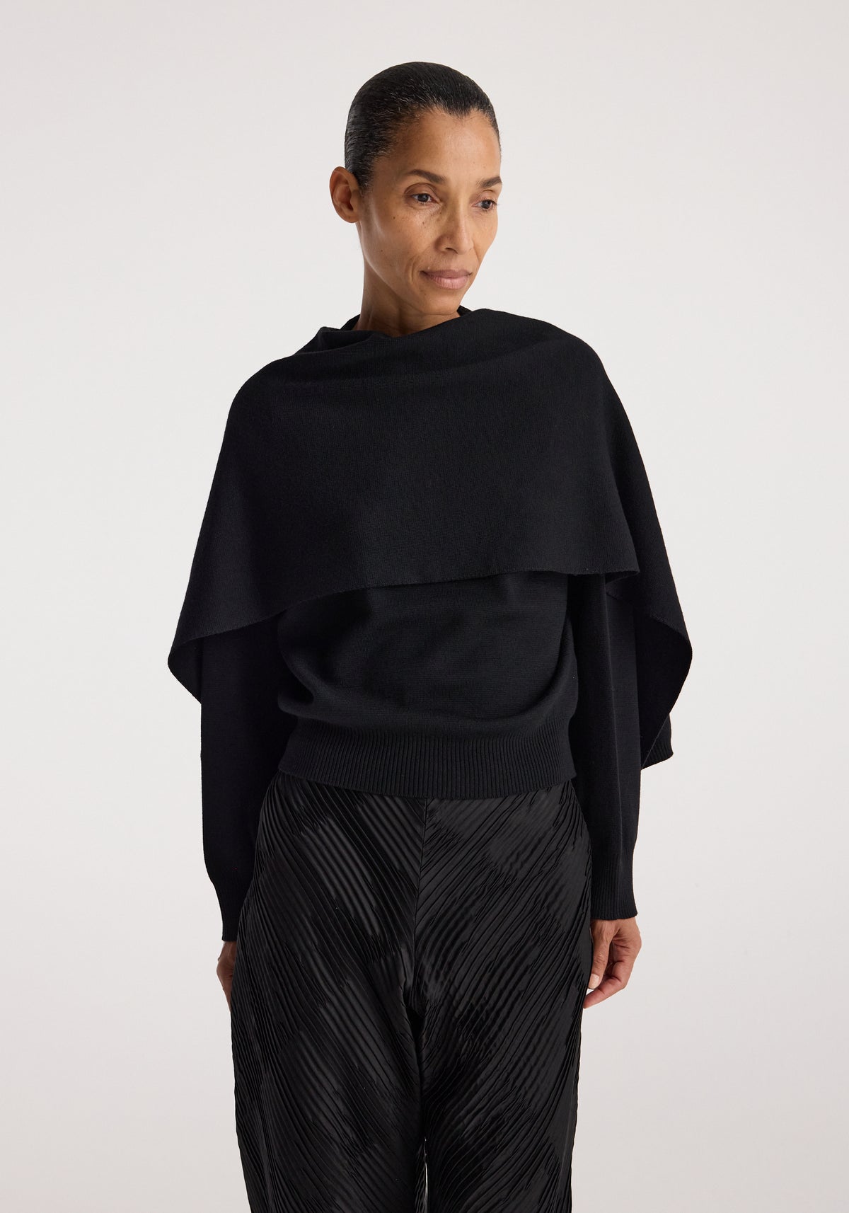 Woman wearing a black Cape jumper | black with a draped cape-like design over the shoulders, paired with textured black pants against a plain background.