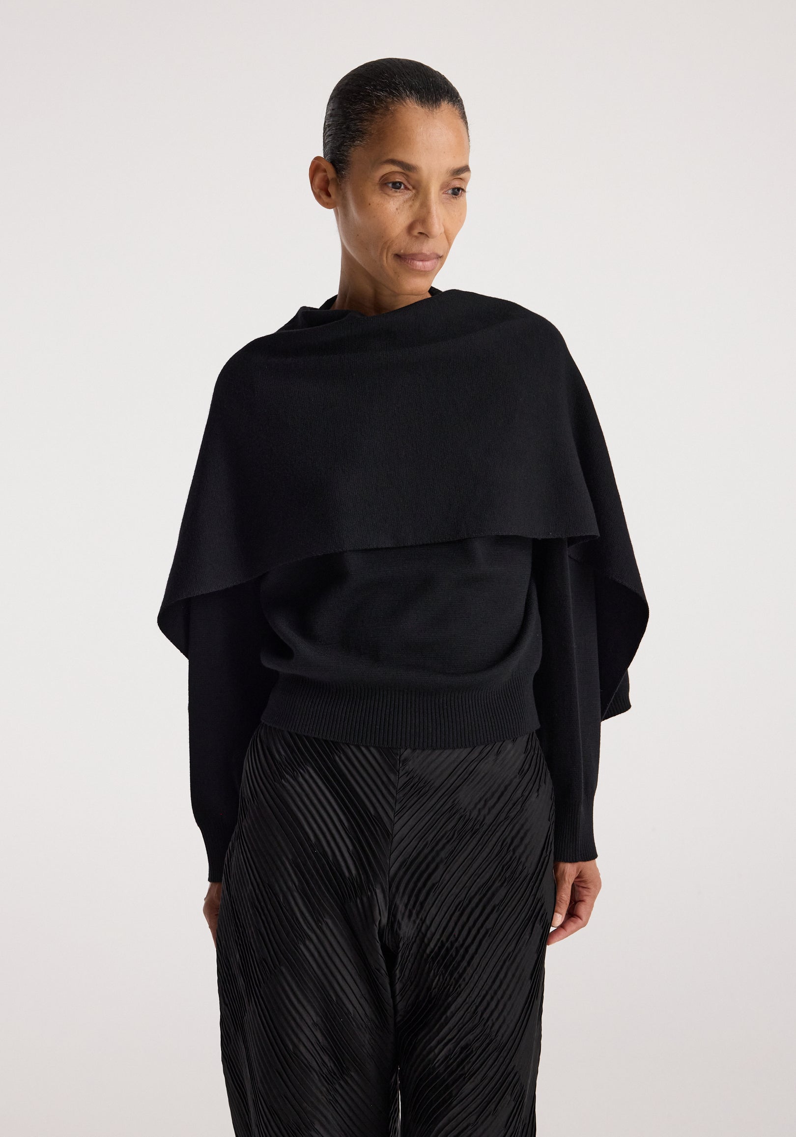 Woman wearing a black Cape jumper | black with a draped cape-like design over the shoulders, paired with textured black pants against a plain background.