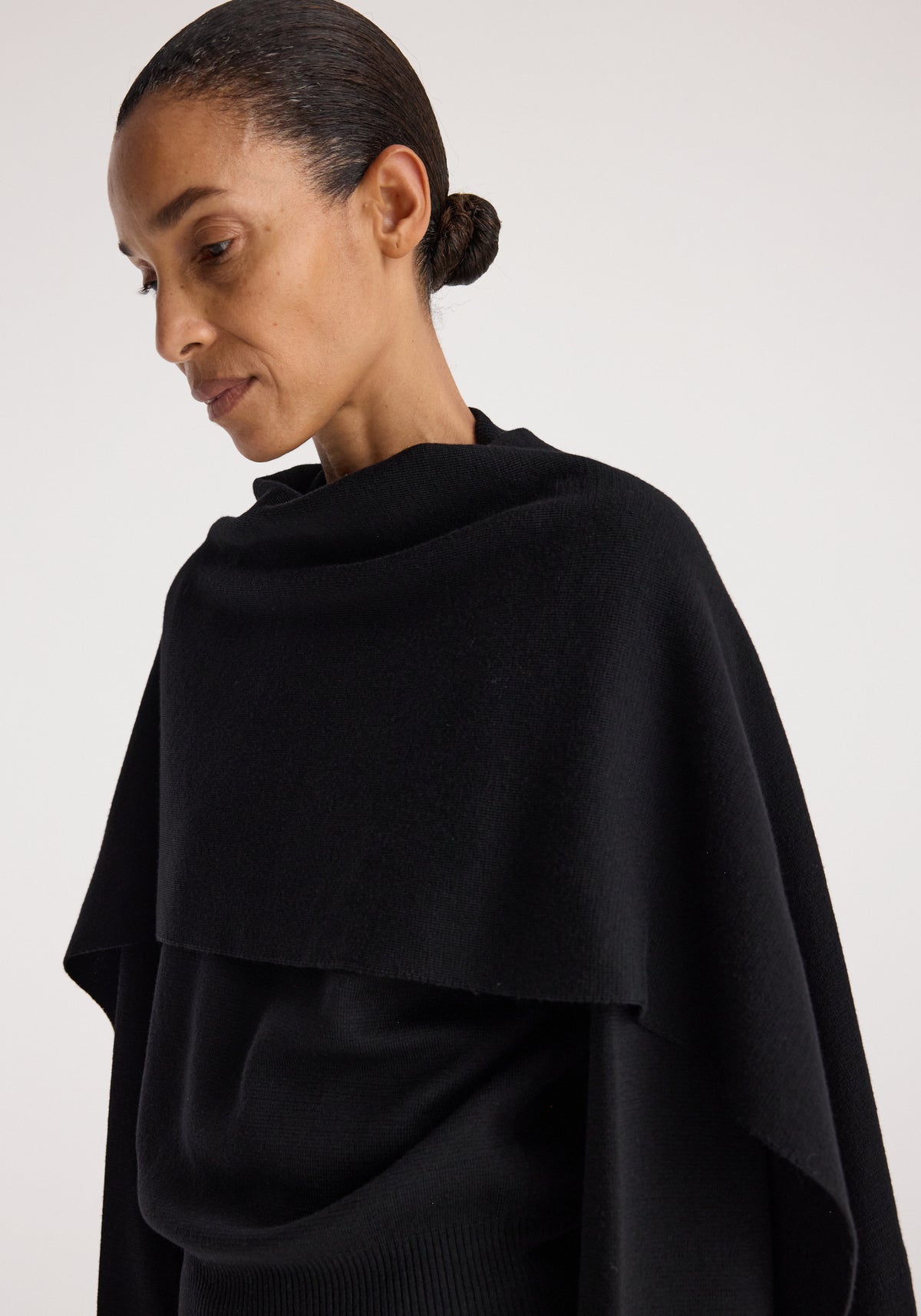 Woman wearing a black Cape jumper | black with a draped, asymmetrical design and a high neckline, styled with hair pulled back in a bun against a plain background.
