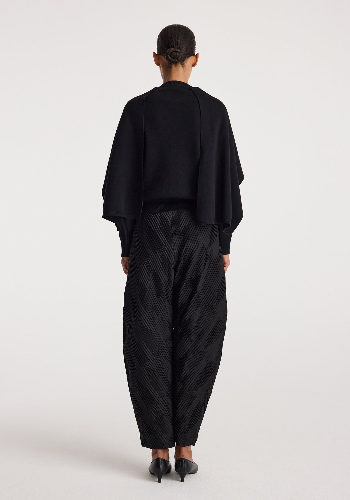 Woman wearing a black Cape jumper | black with wide, textured black pants and black heeled shoes, shown from the back against a plain background.