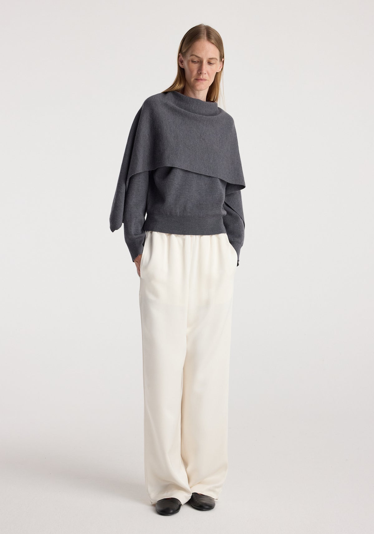 Woman wearing a dark grey melange knitted Cape jumper | dark grey melange paired with loose white trousers and black shoes standing against a plain background.