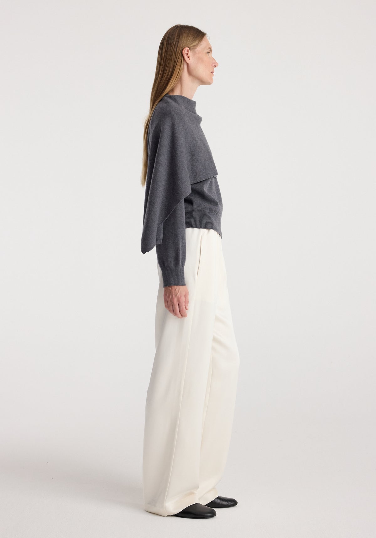 Side view of a woman wearing a dark grey melange Cape jumper | dark grey melange paired with white wide-leg trousers and black shoes against a plain background.
