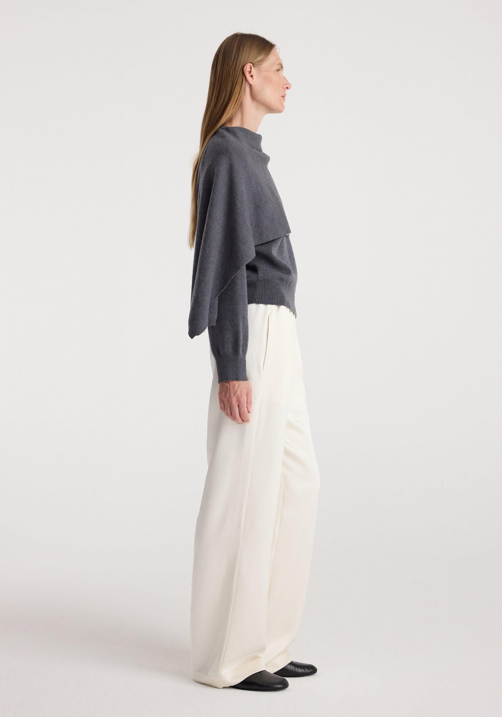 Side view of a woman wearing a dark grey melange Cape jumper | dark grey melange paired with white wide-leg trousers and black shoes against a plain background.