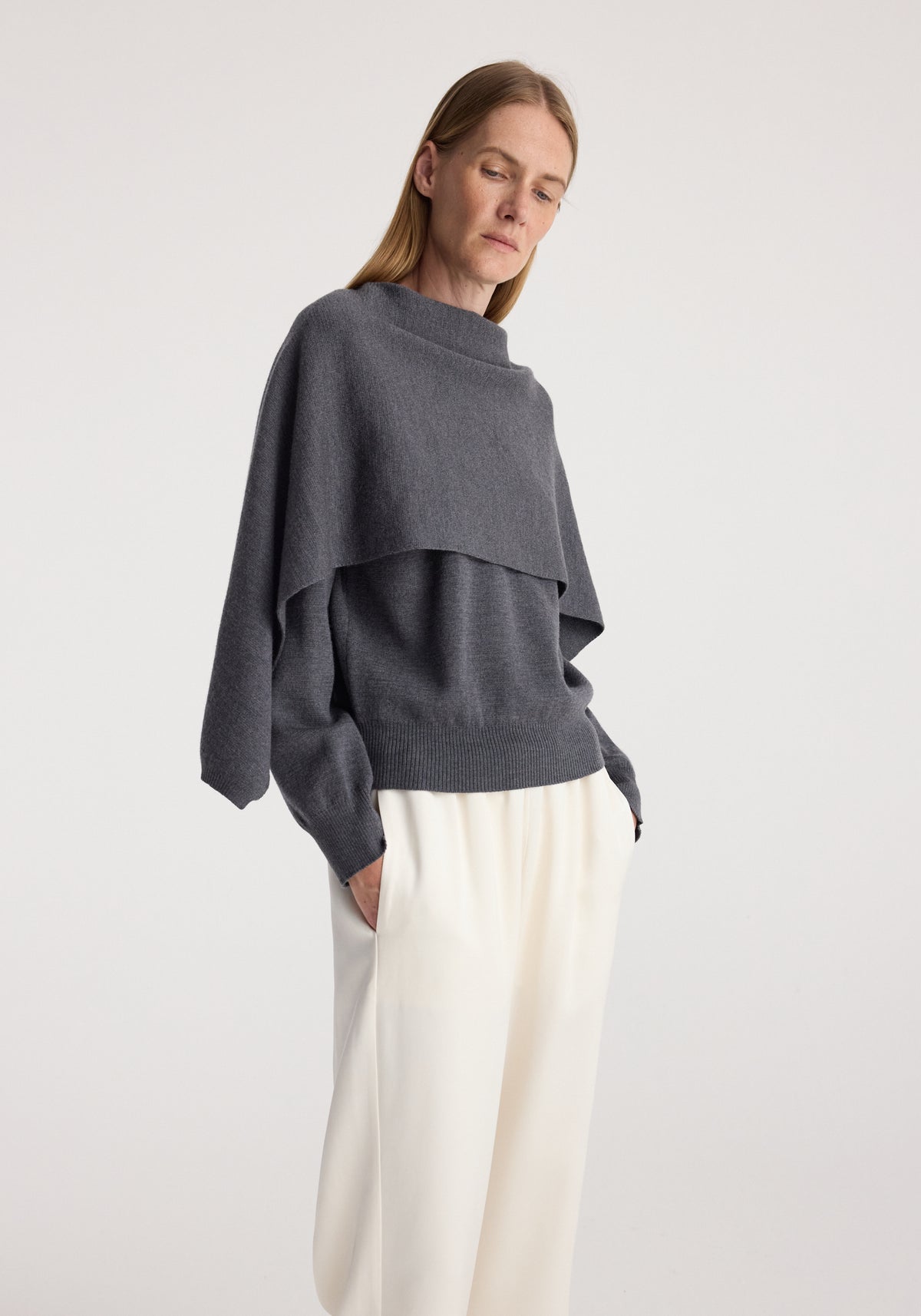 Woman wearing a dark grey melange layered sweater with long sleeves and a high neckline paired with loose white pants, showcasing the Cape jumper | dark grey melange.