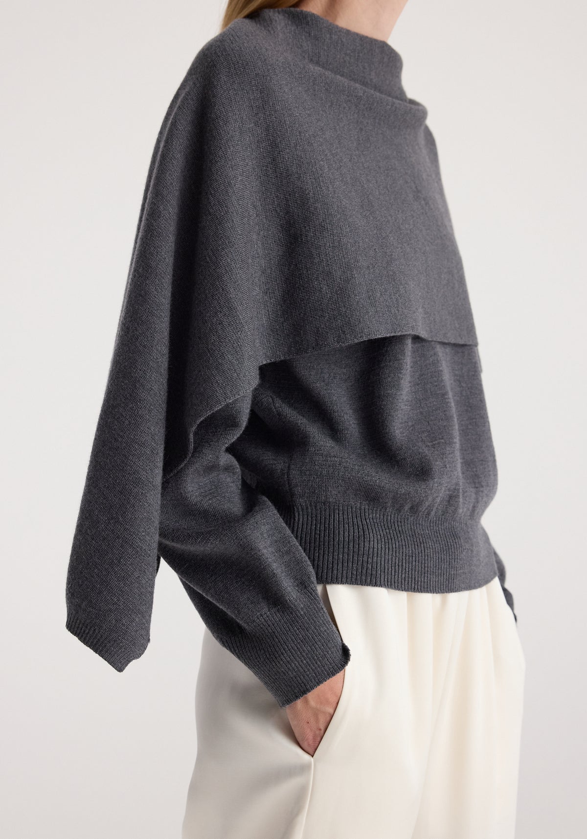 Side view of a person wearing a dark grey melange layered knit sweater with long sleeves and a high neckline paired with cream-colored pants, showcasing the Cape jumper | dark grey melange.