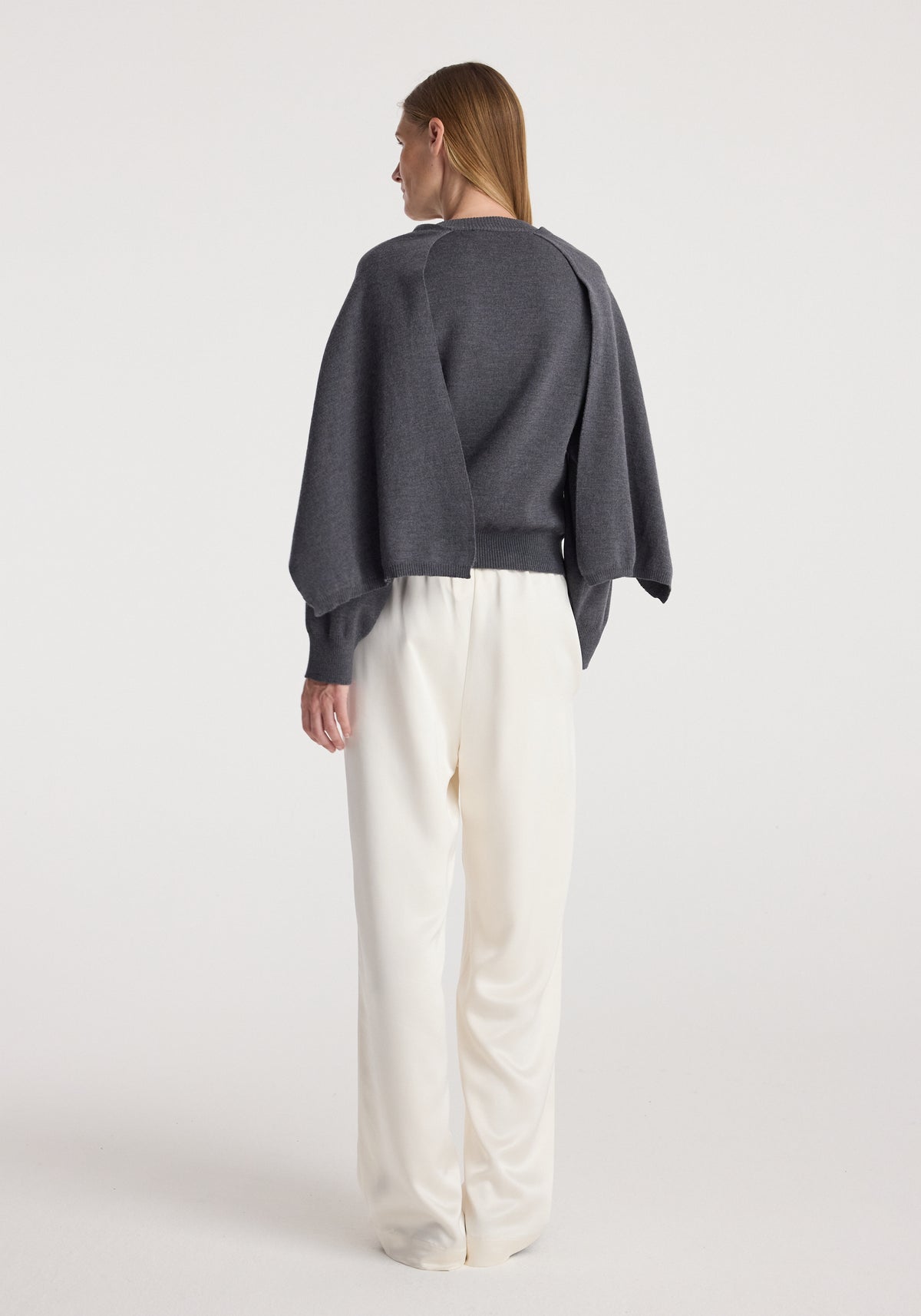 Woman seen from the back wearing a Cape jumper | dark grey melange paired with loose white pants against a plain light background.
