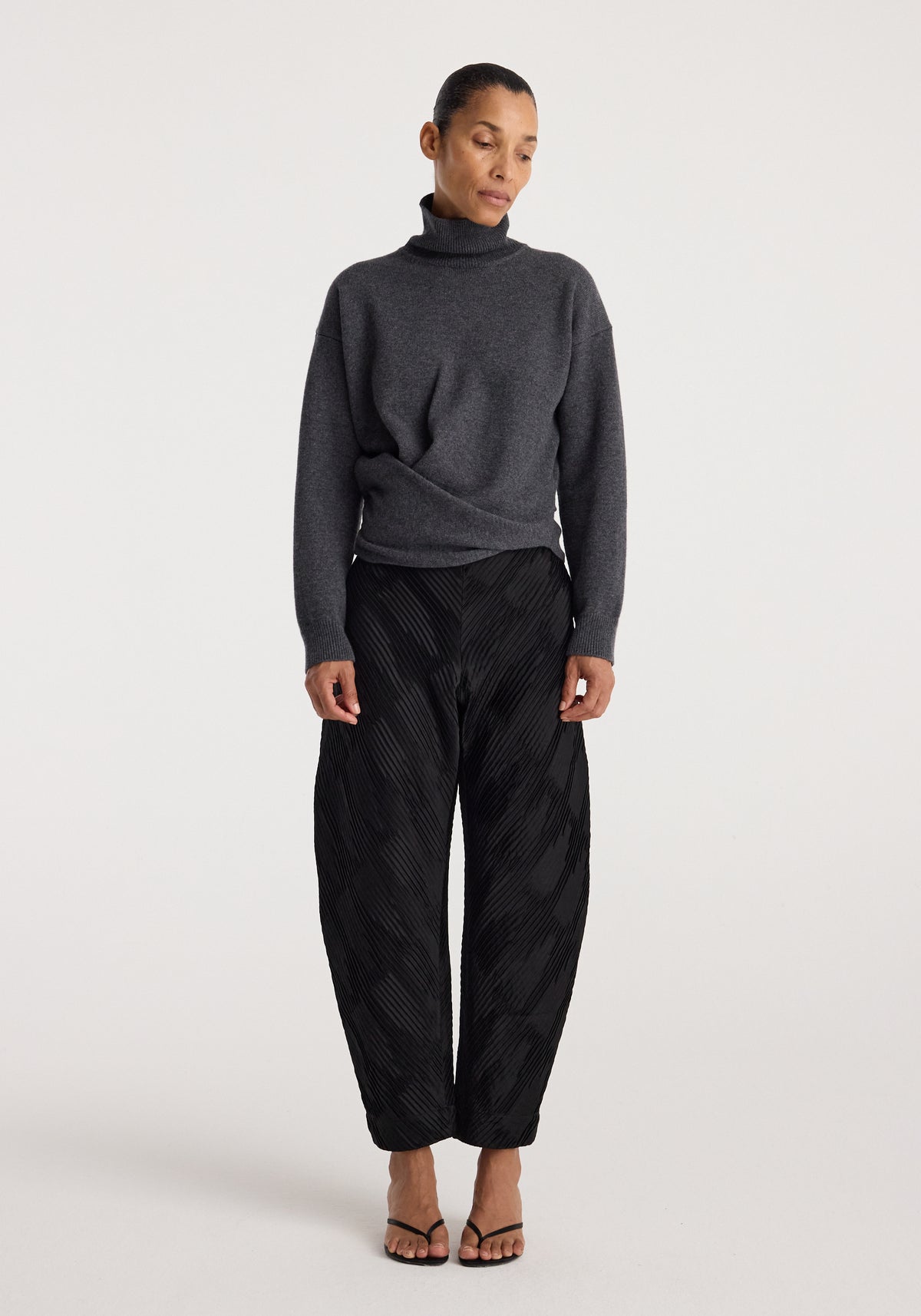 Woman wearing a Sculptural turtleneck | dark grey melange paired with black wide-leg textured pants and black open-toe heels, standing against a plain background.