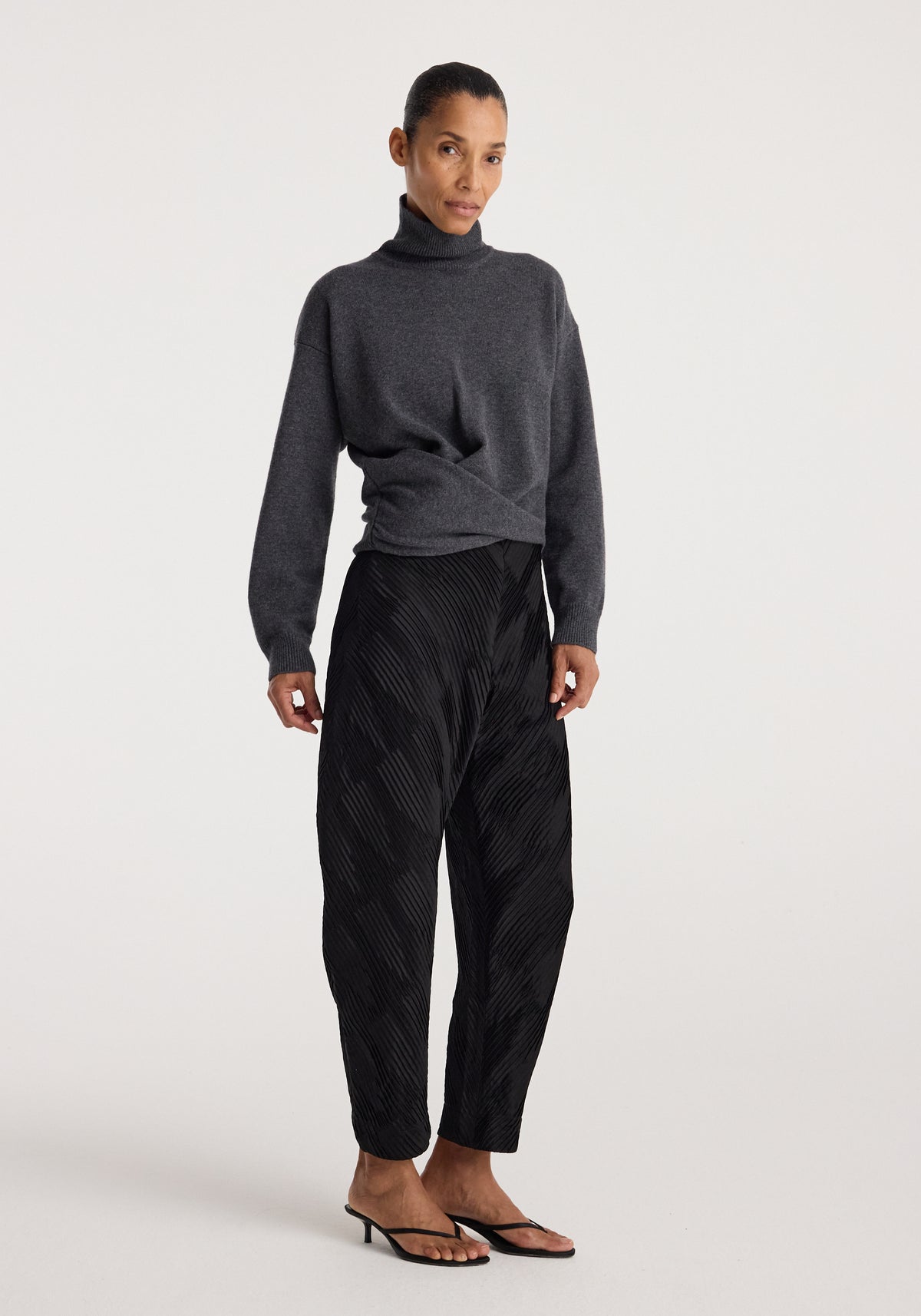 Woman wearing a Sculptural turtleneck | dark grey melange with long sleeves, paired with black patterned pants and black heeled sandals, standing against a white background.