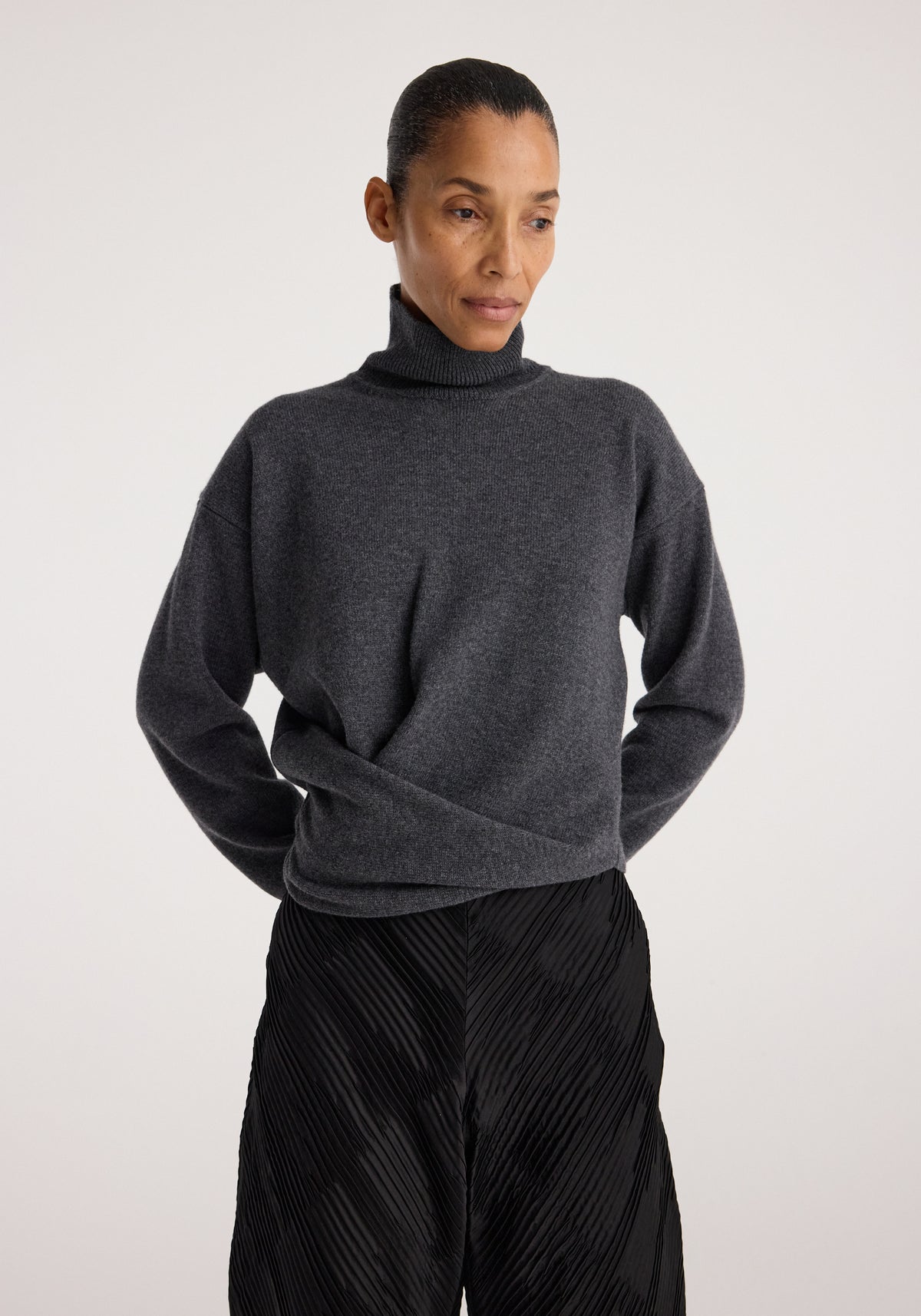 Woman wearing a Sculptural turtleneck | dark grey melange sweater paired with textured black wide-leg pants against a plain background