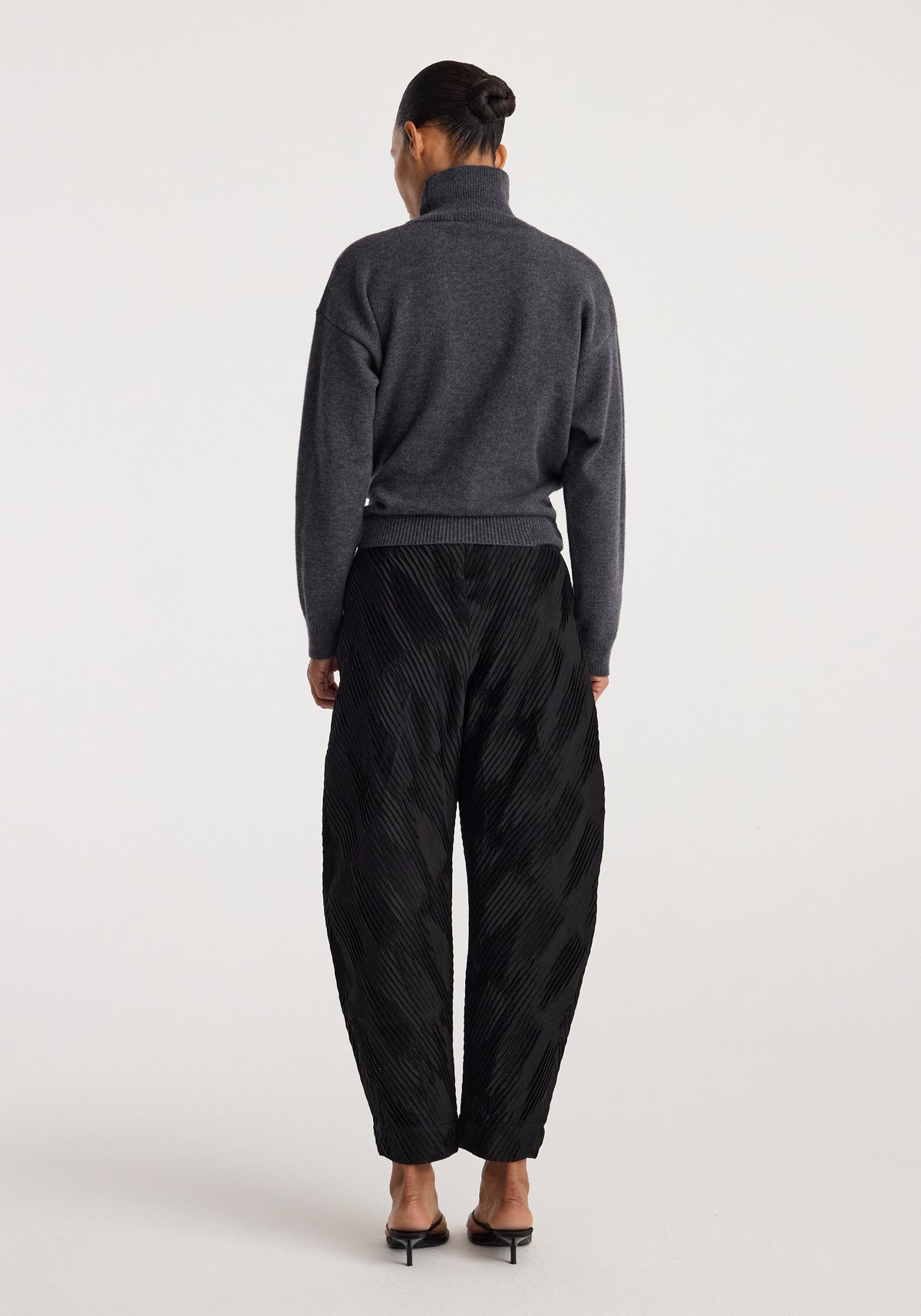 Model wearing a Sculptural turtleneck | dark grey melange with long sleeves, standing facing away, paired with black textured wide-leg pants and black heeled sandals.