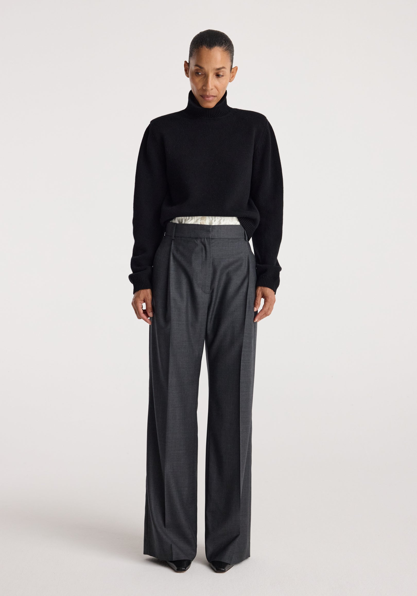 Woman wearing a black Statement shoulder turtleneck | black sweater paired with high-waisted dark gray wide-leg trousers against a plain light background.