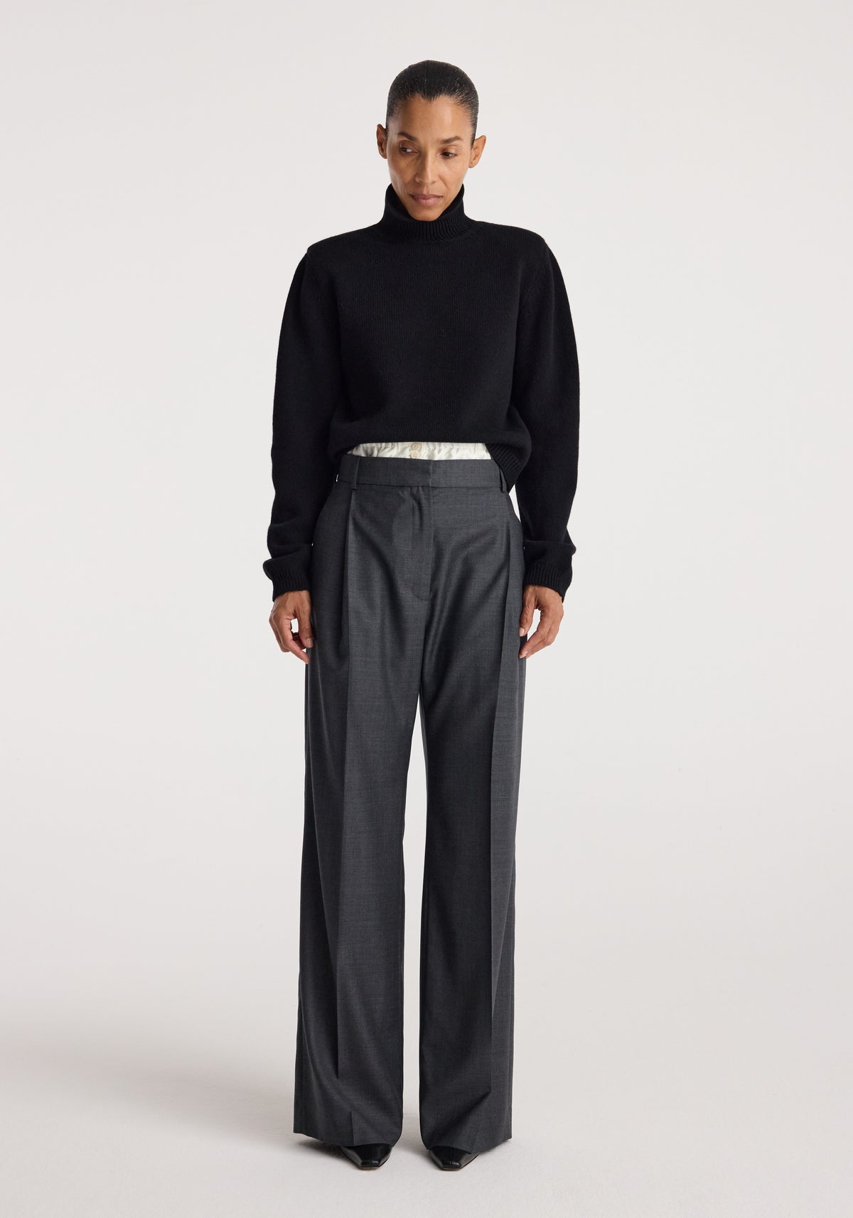 Woman wearing a black Statement shoulder turtleneck | black sweater paired with high-waisted dark gray wide-leg trousers against a plain light background.