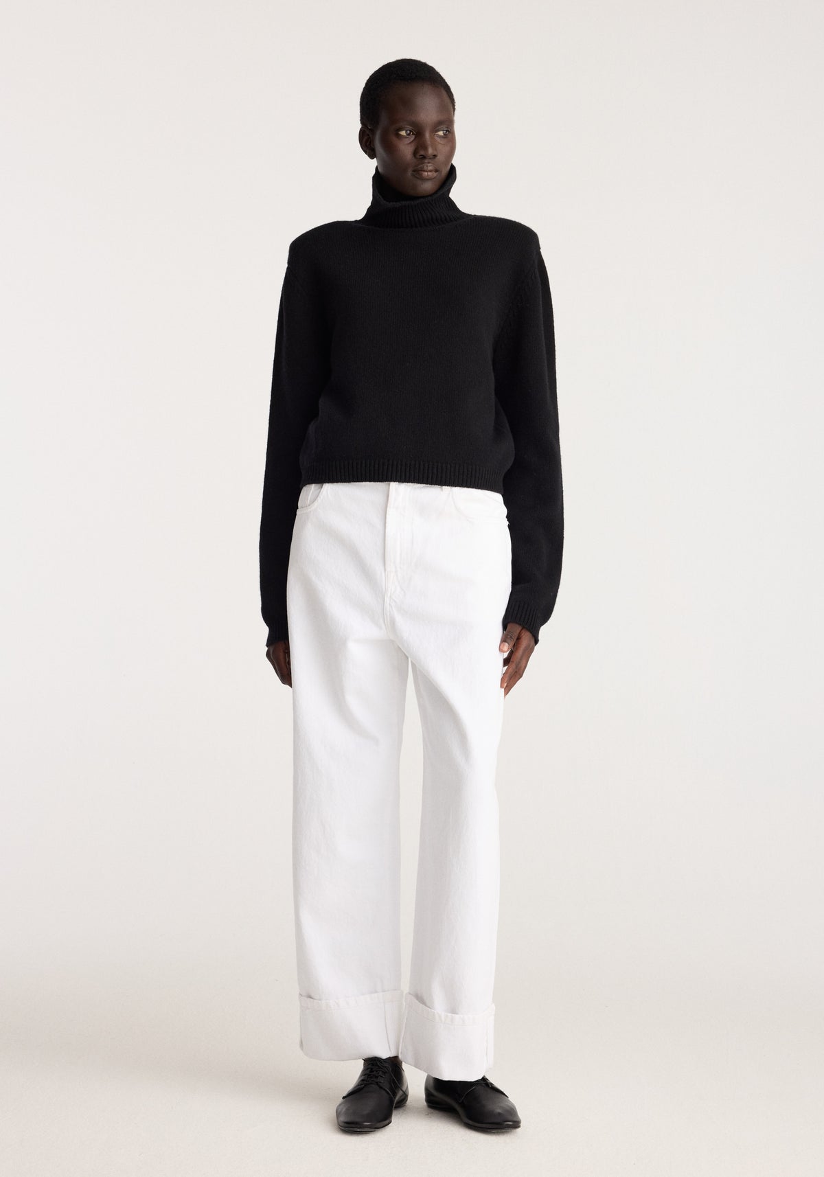 Statement shoulder turtleneck | black worn with white cuffed trousers and black shoes on a model against a neutral studio background
