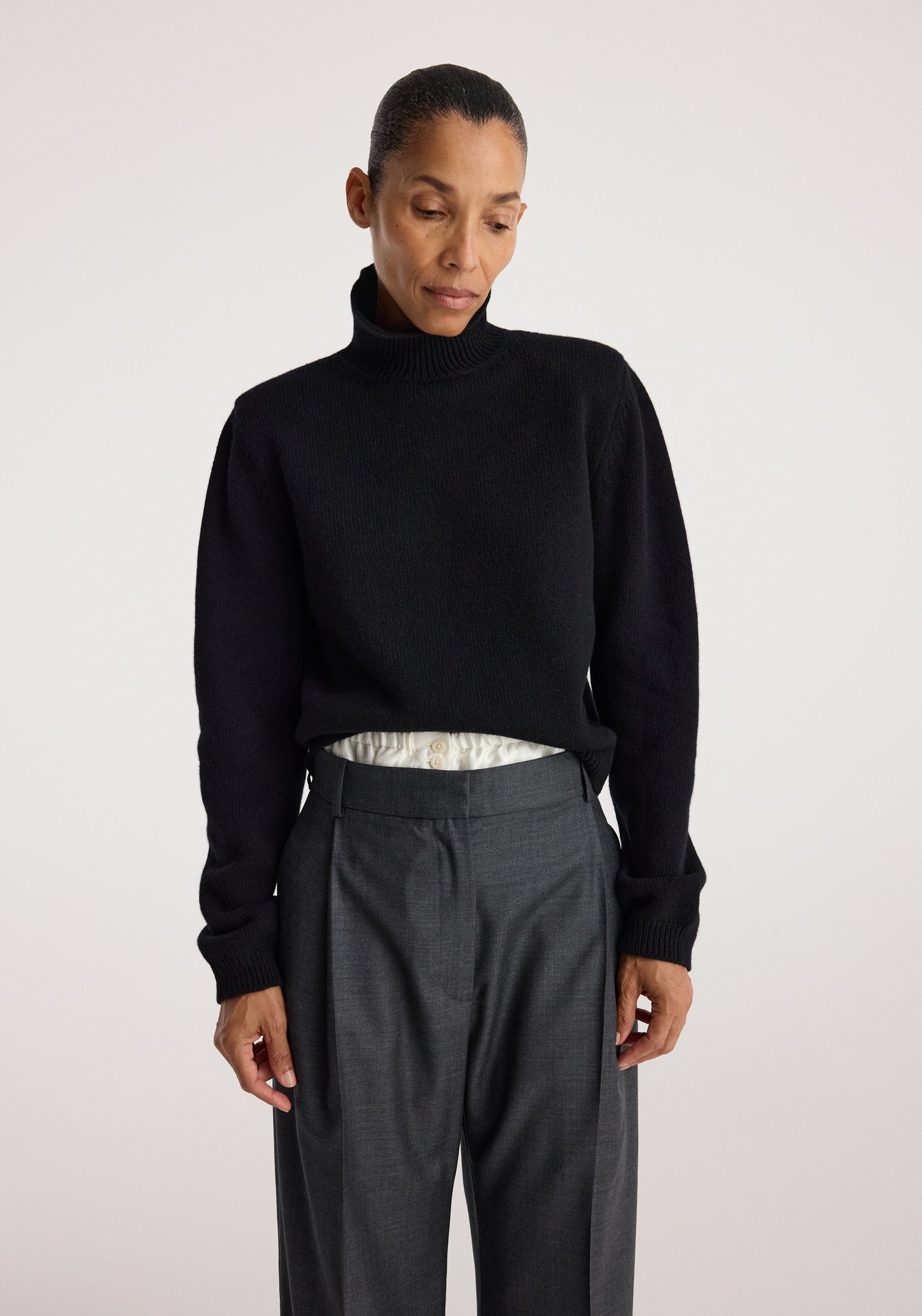 Woman wearing a black Statement shoulder turtleneck | black with long sleeves, paired with high-waisted gray trousers against a plain background.