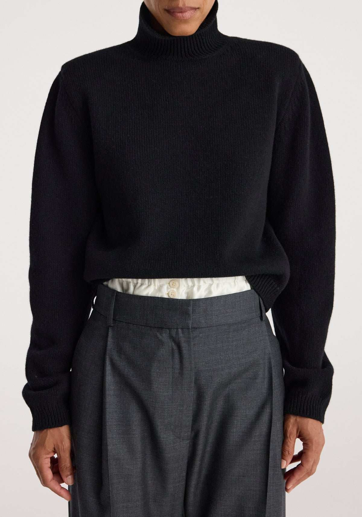 Close-up of a model wearing a black Statement shoulder turtleneck | black paired with high-waisted gray trousers and a tucked-in white shirt showing button details.