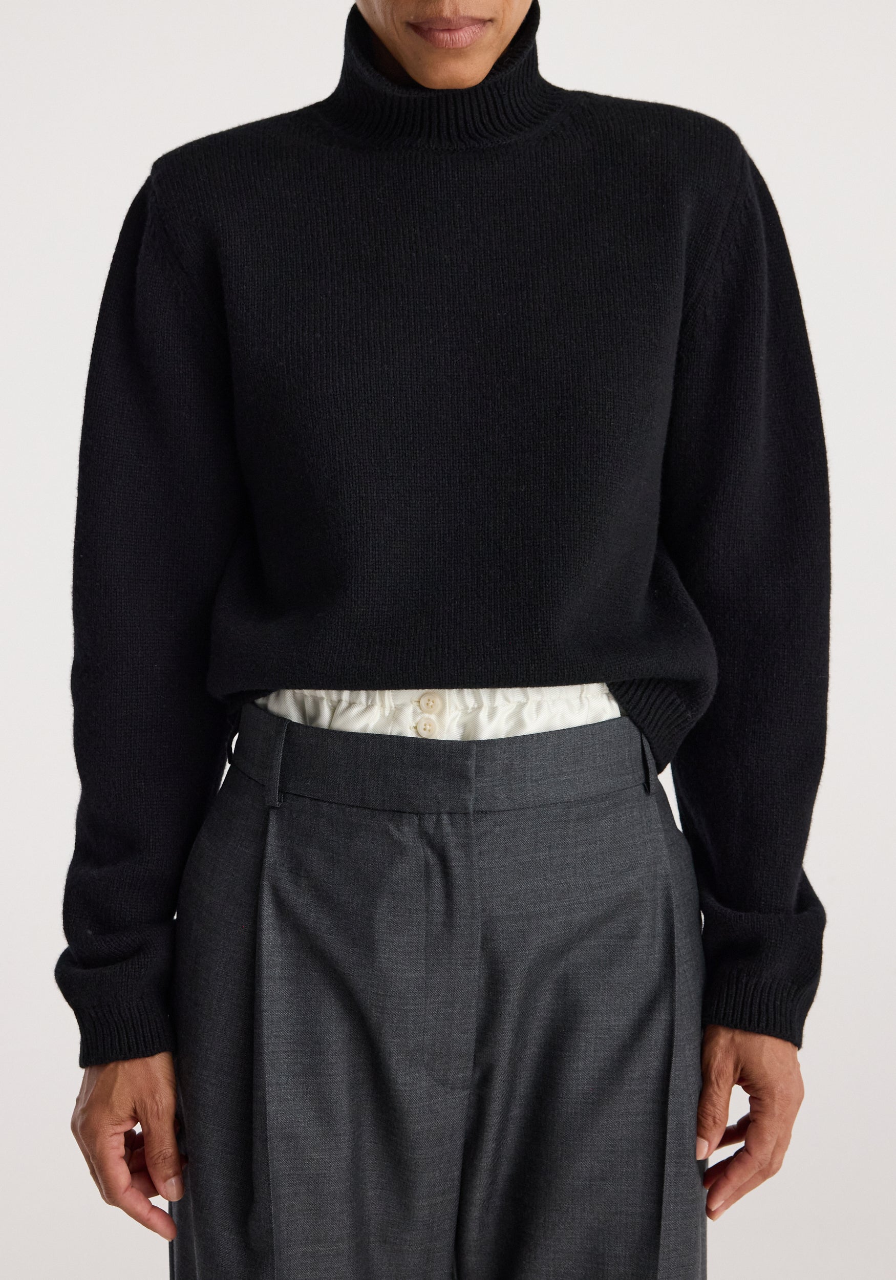 Close-up of a model wearing a black Statement shoulder turtleneck | black paired with high-waisted gray trousers and a tucked-in white shirt showing button details.
