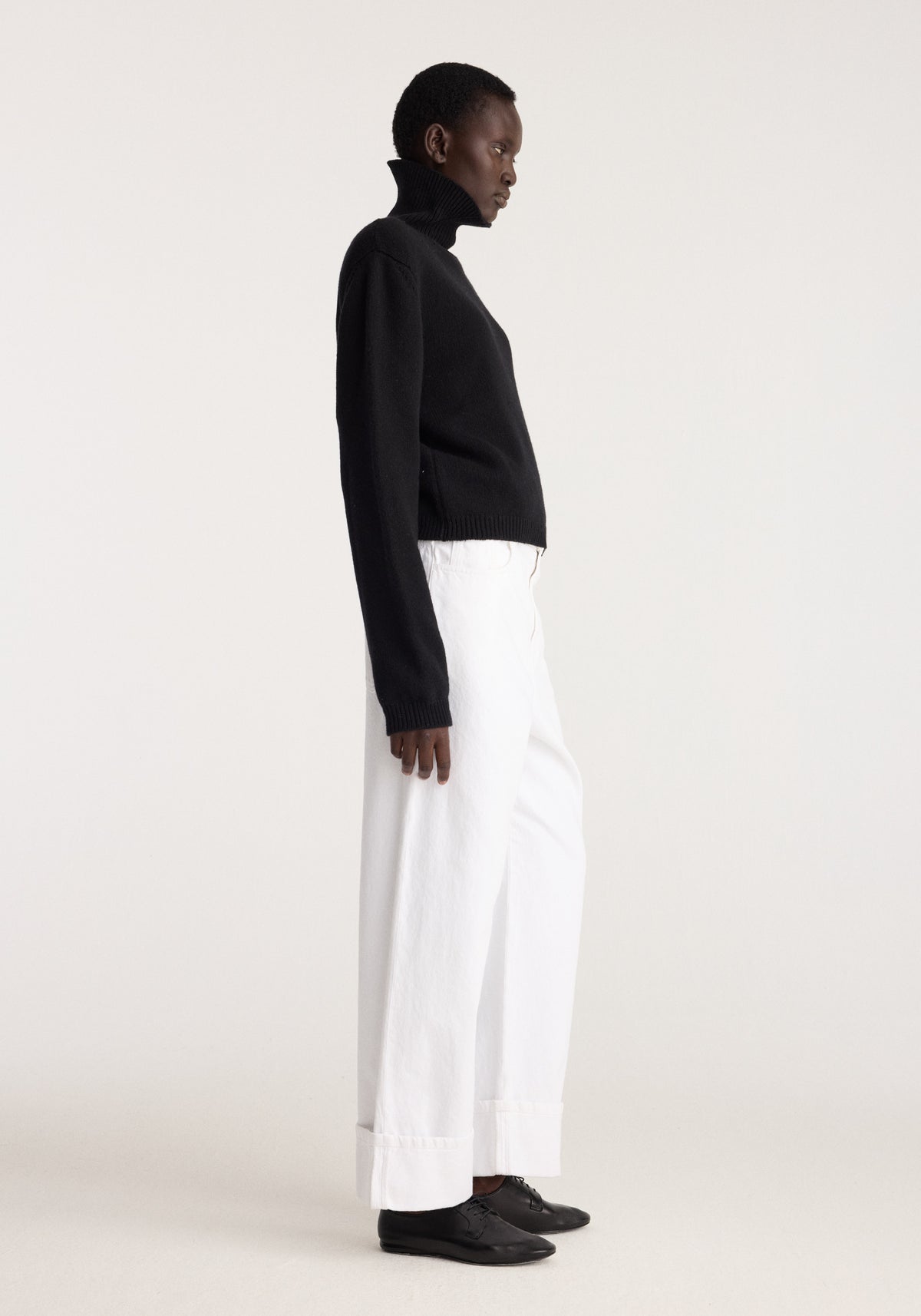 Statement shoulder turtleneck | black worn with white wide leg trousers and black shoes on a model in side profile against a neutral background