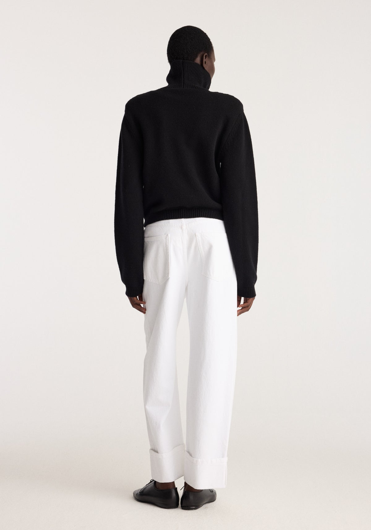Back view of a model wearing the Statement shoulder turtleneck | black with white trousers and black shoes in a minimalist studio setting