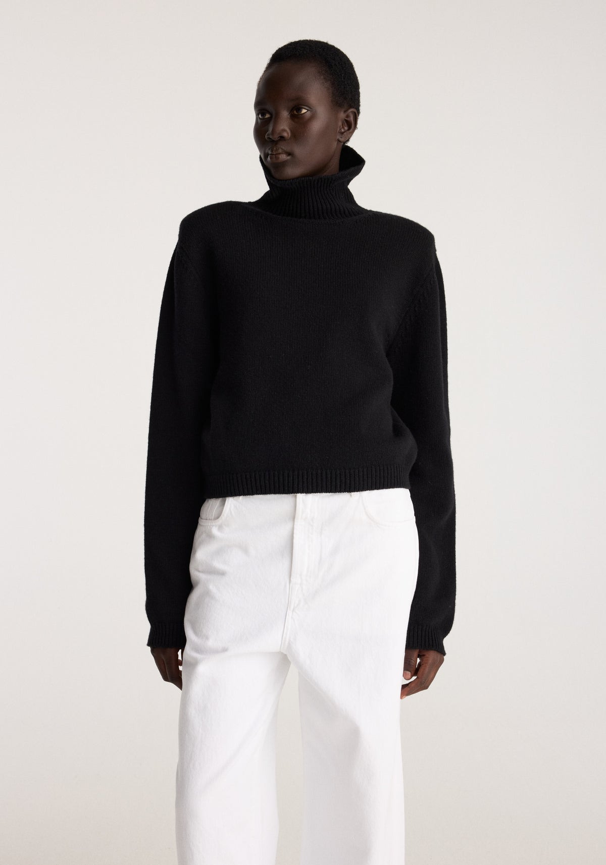 Statement shoulder turtleneck | black worn with white trousers by a model against a light neutral background