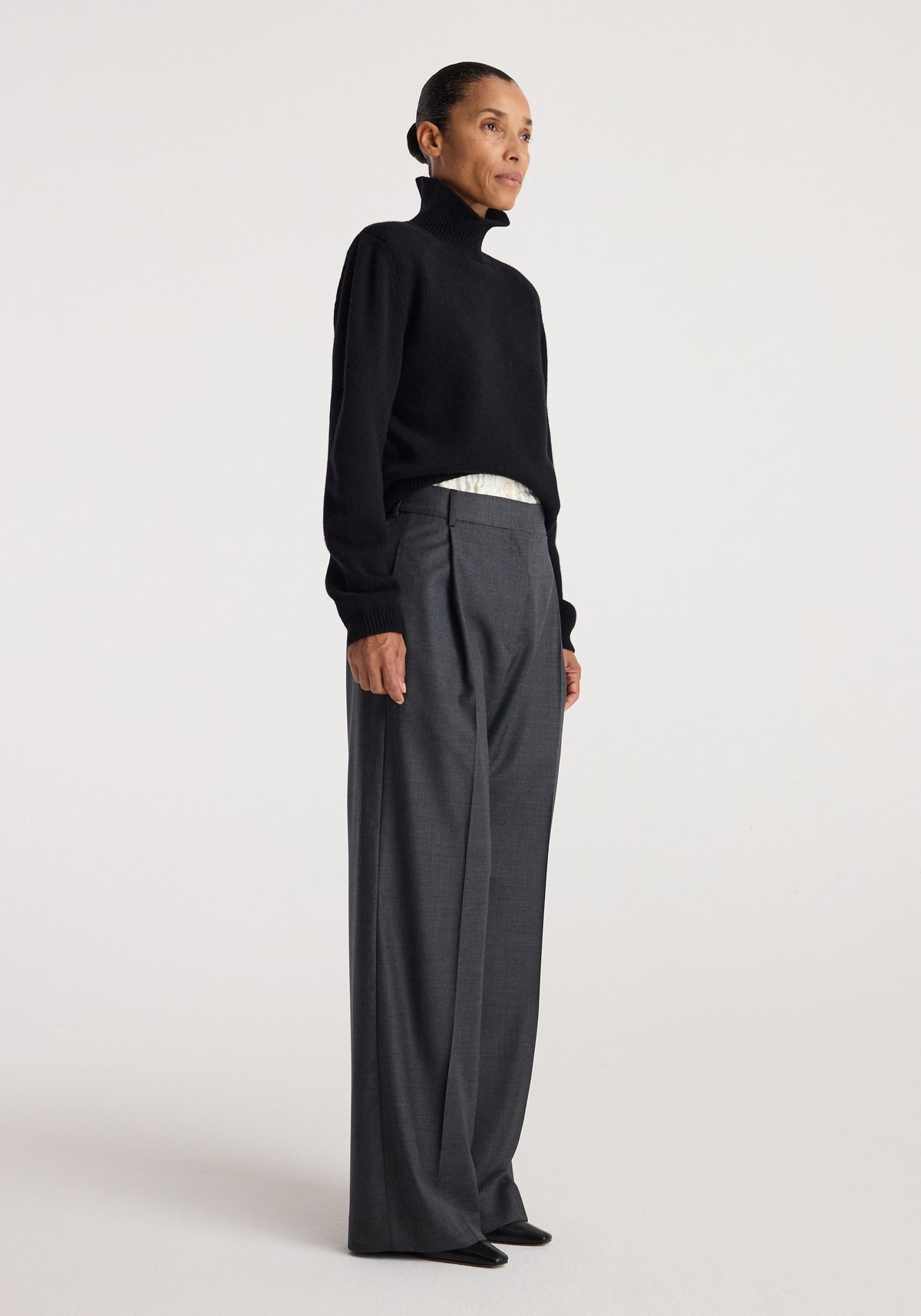 Woman standing in a black Statement shoulder turtleneck | black paired with high-waisted wide-leg gray trousers against a plain background.