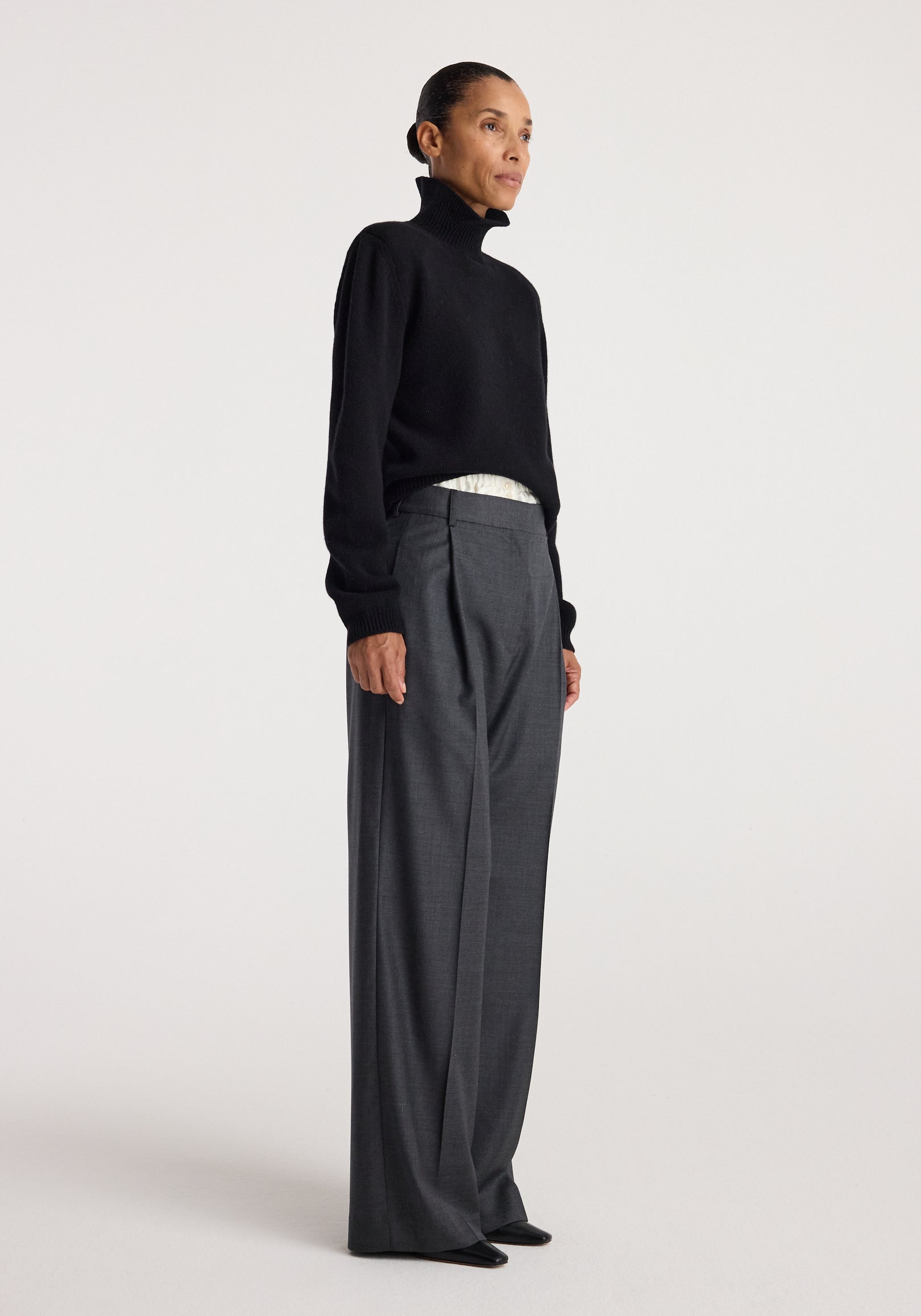 Woman standing in a black Statement shoulder turtleneck | black paired with high-waisted wide-leg gray trousers against a plain background.