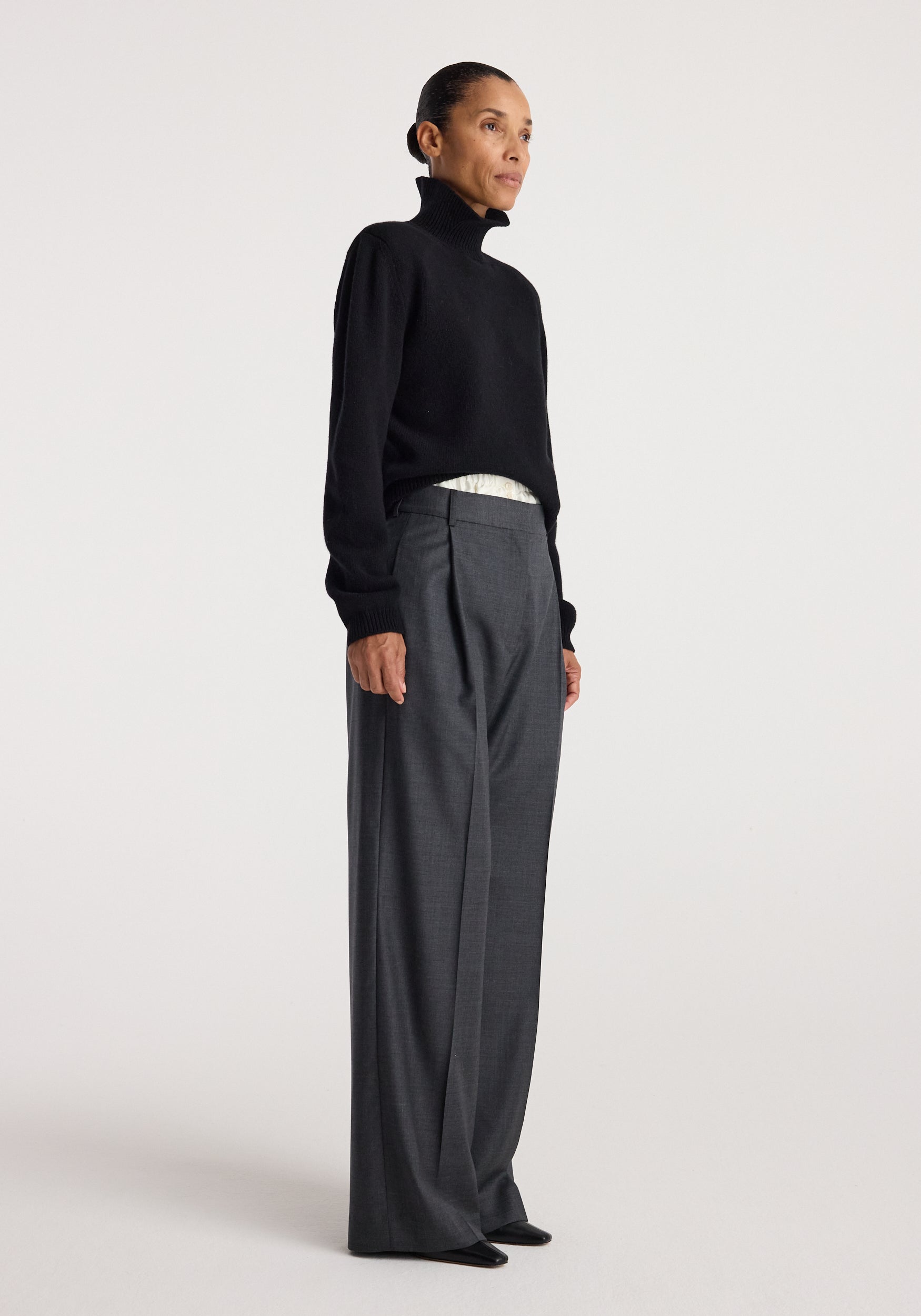 Woman standing in a black Statement shoulder turtleneck | black paired with high-waisted wide-leg gray trousers against a plain background.