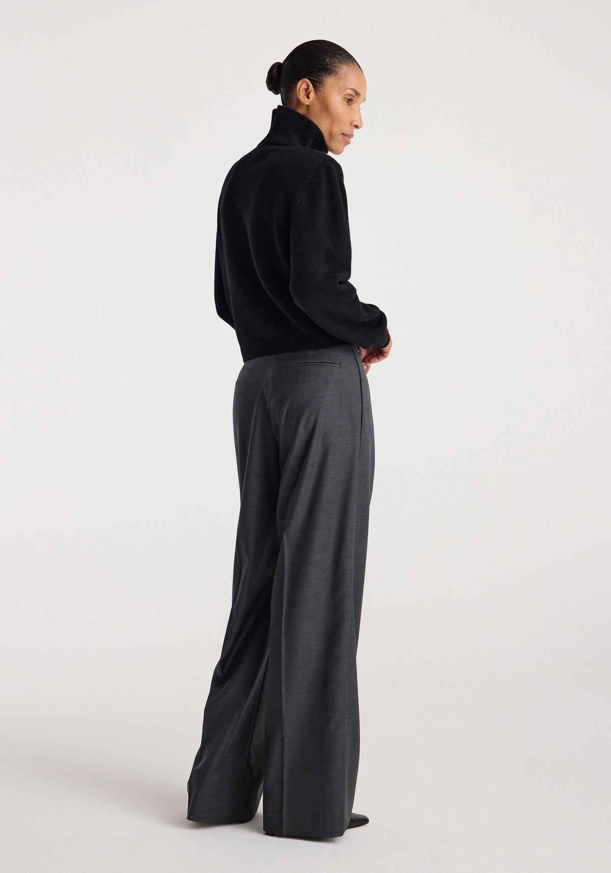 Woman wearing a black Statement shoulder turtleneck | black with long sleeves, paired with high-waisted gray wide-leg pants, standing in profile against a plain background.