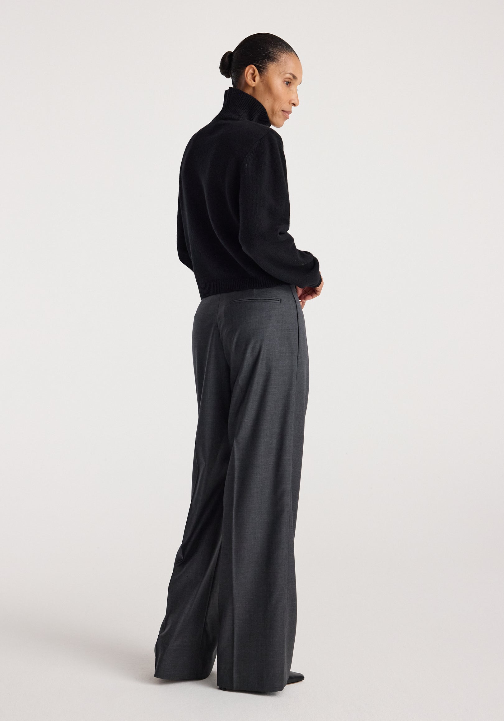 Woman wearing a black Statement shoulder turtleneck | black with long sleeves, paired with high-waisted gray wide-leg pants, standing in profile against a plain background.