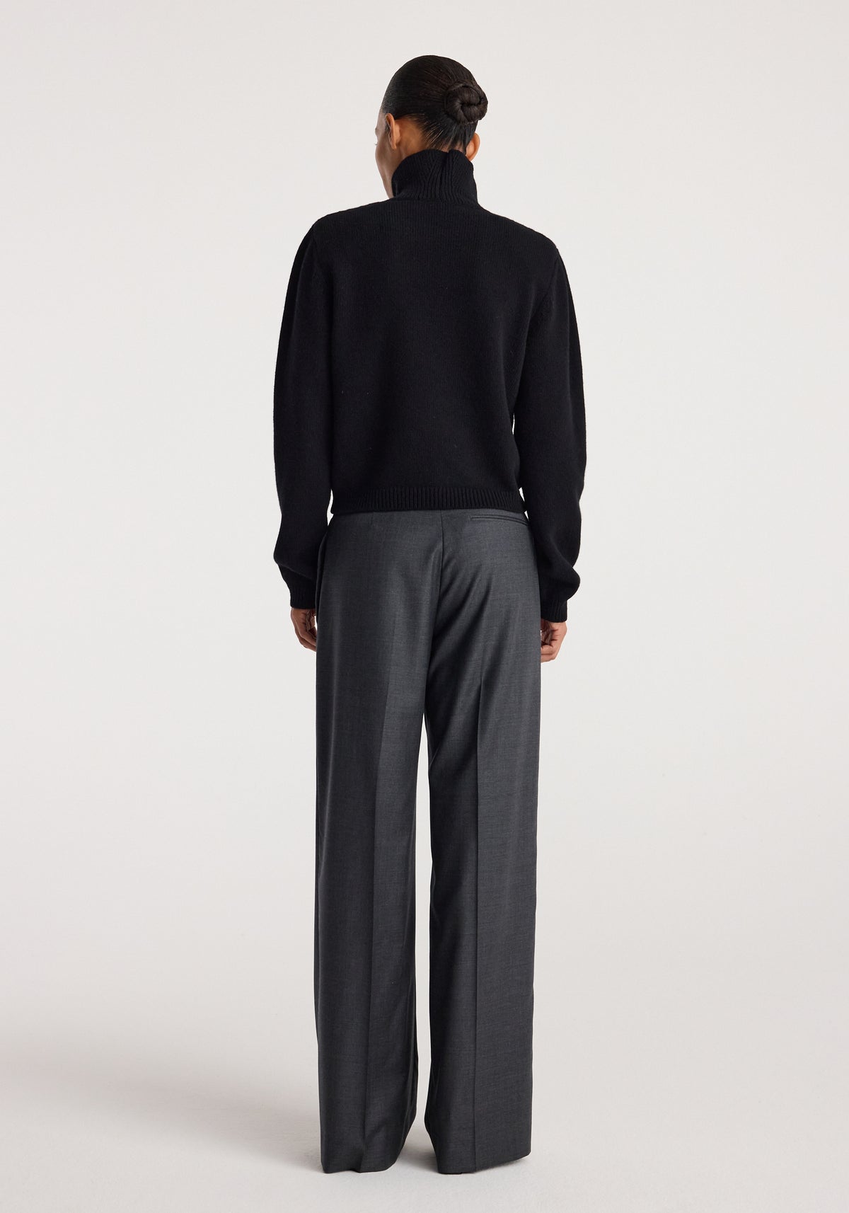 Back view of a person wearing the Statement shoulder turtleneck | black with long sleeves and a high collar, paired with loose dark gray trousers against a plain background.