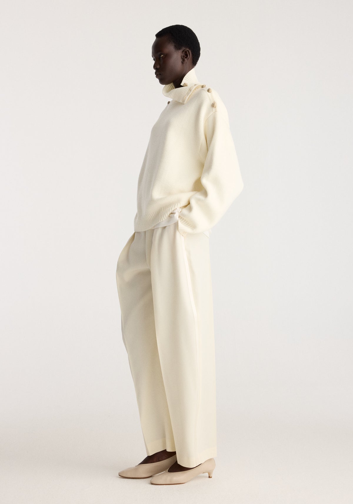 Uneven sailor polo | cream worn by a model in a minimalist studio with relaxed cream trousers and pointed flats posing in profile