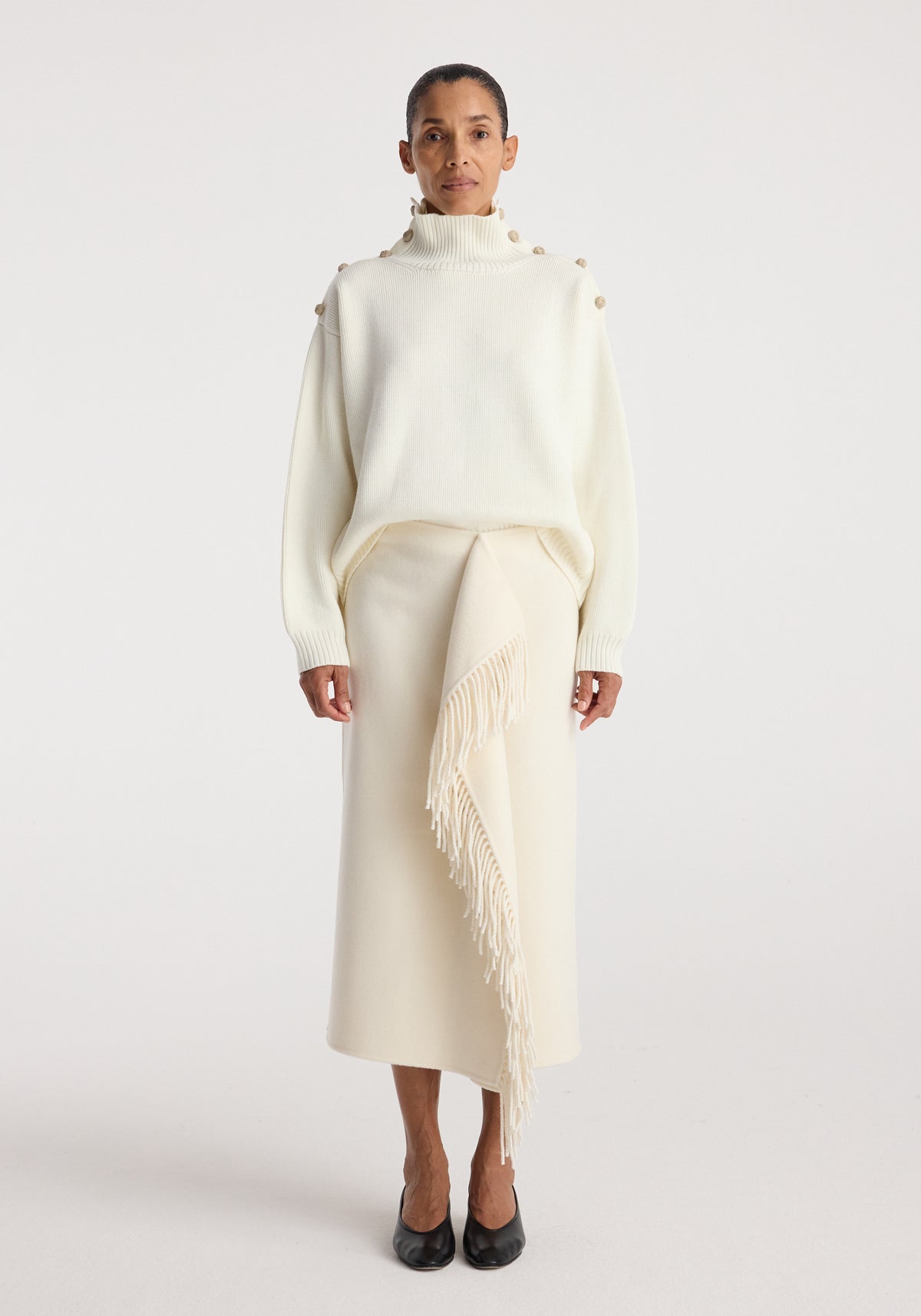 Woman wearing an oversized cream sweater with button details along the shoulders and a high neck, paired with a cream skirt featuring a long fringed panel, styled with black shoes, showcasing the Uneven sailor polo | cream.