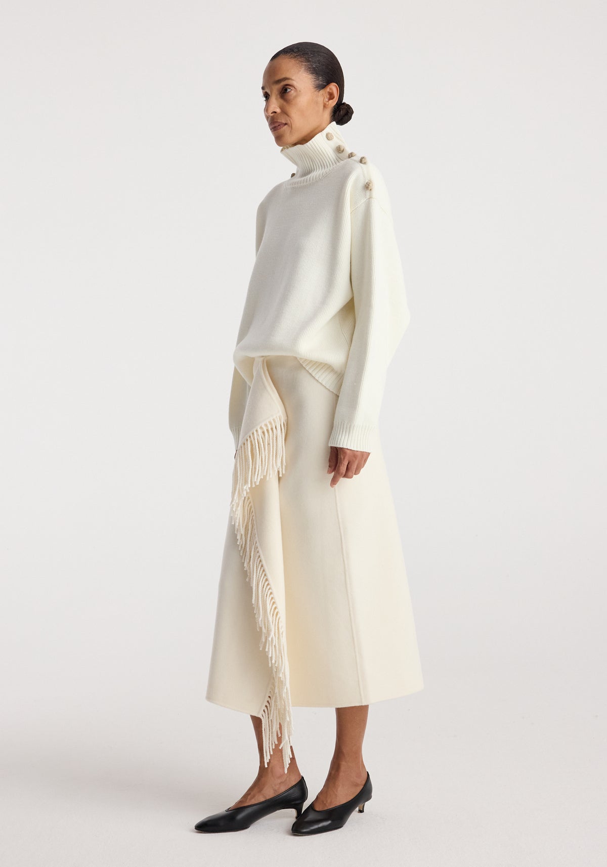 Woman wearing an uneven sailor polo | cream with button details on the shoulder paired with a cream midi skirt featuring long fringe accents, and black low-heeled shoes.