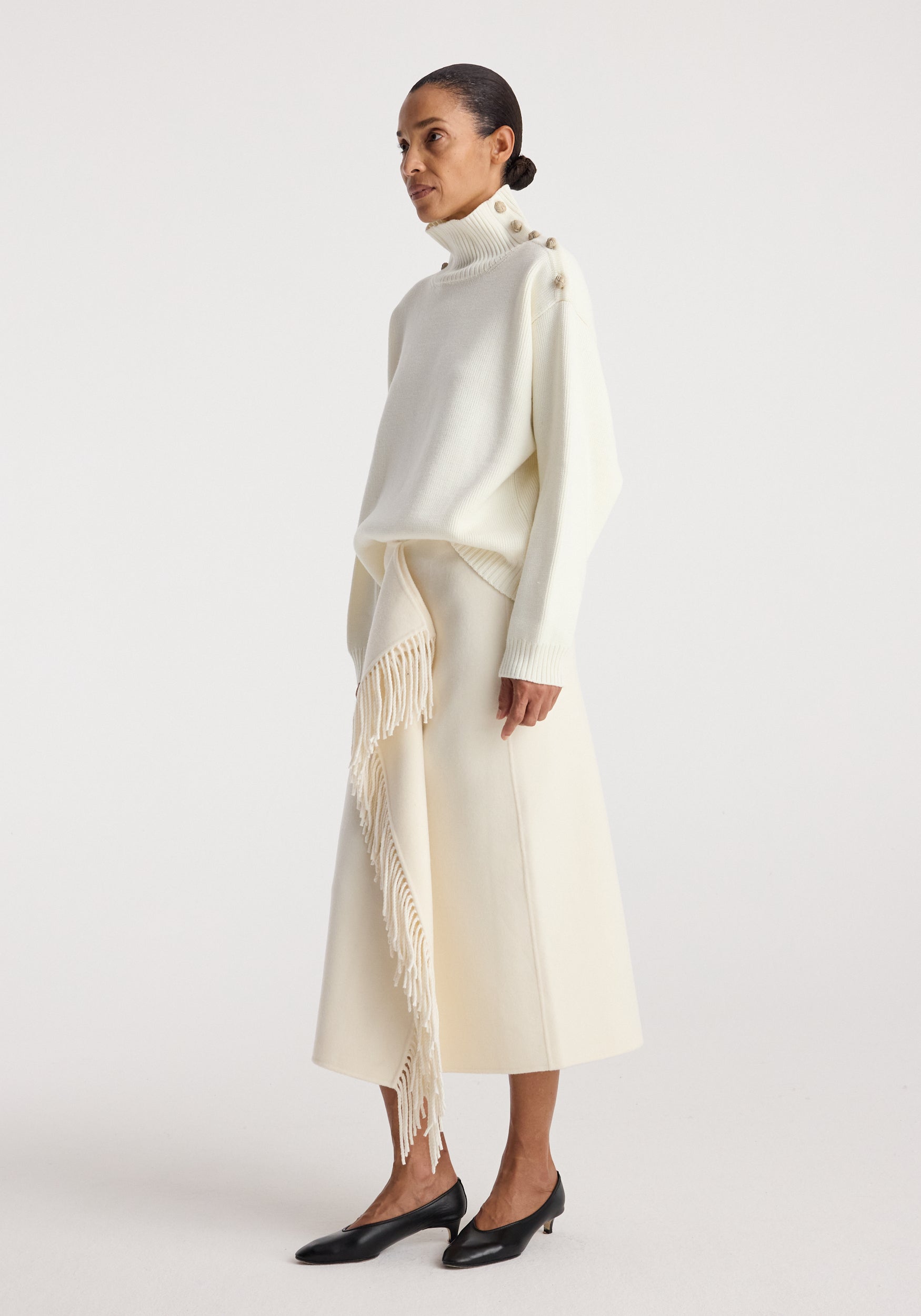 Woman wearing an uneven sailor polo | cream with button details on the shoulder paired with a cream midi skirt featuring long fringe accents, and black low-heeled shoes.