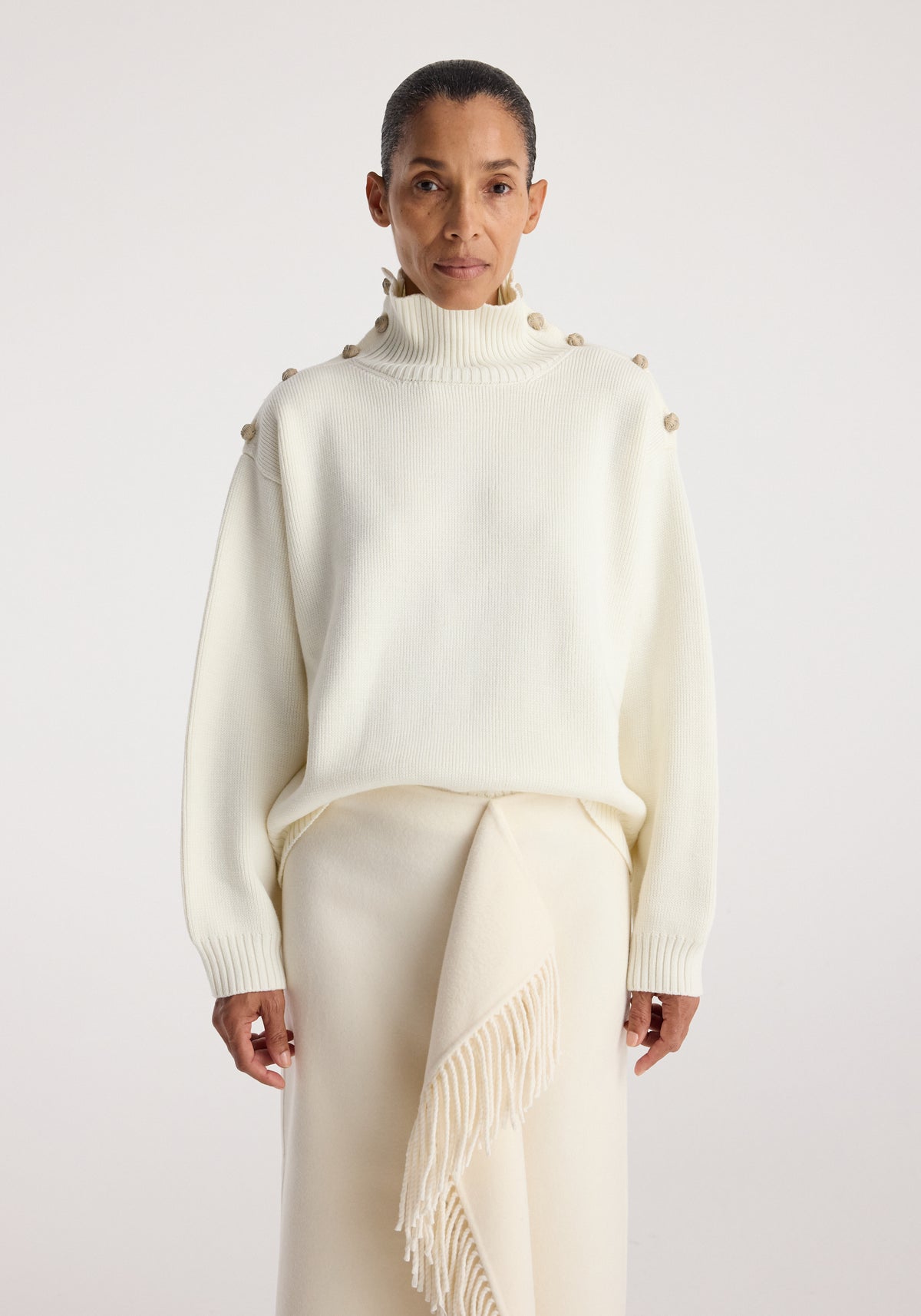Woman wearing an uneven sailor polo | cream with button details on the shoulders and a high neckline, styled with a matching cream skirt featuring a fringed detail.