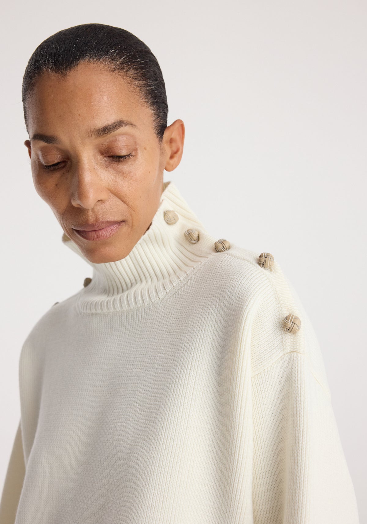 Woman wearing a cream knit sweater with a high, ribbed collar featuring button details along the shoulder in the Uneven sailor polo | cream style.