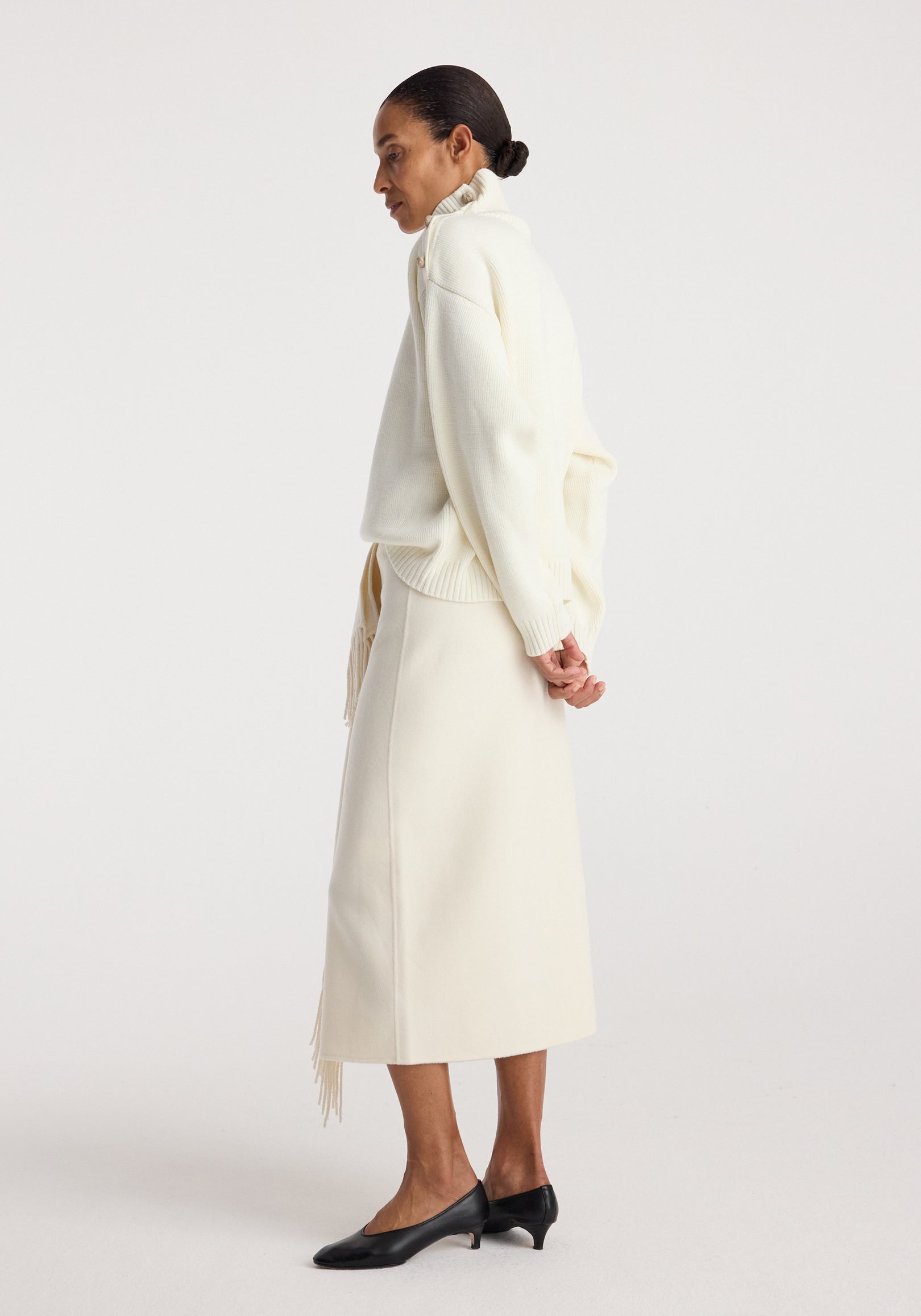 Side view of a woman wearing an uneven sailor polo | cream with long sleeves and a high collar, paired with a matching cream skirt and black low-heeled shoes.