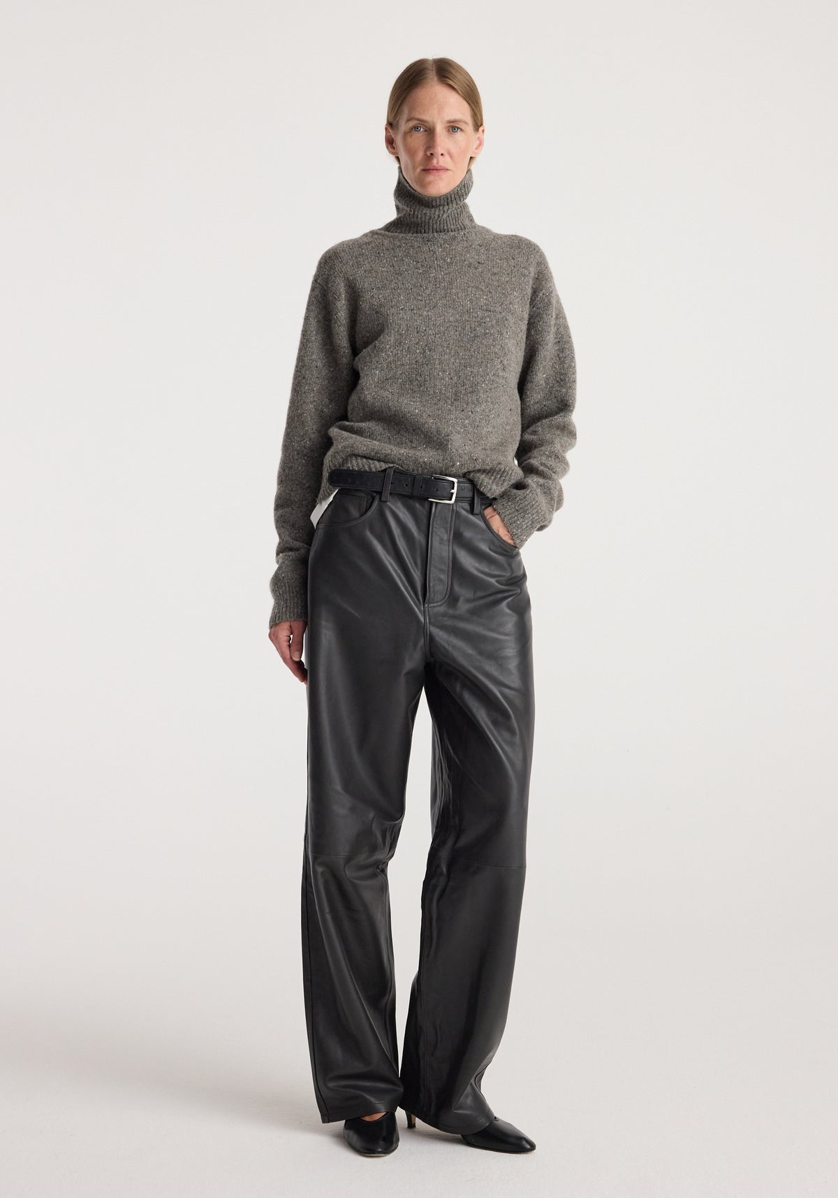 Woman wearing a grey mouliné turtleneck with long sleeves, tucked into high-waisted black leather pants with a black belt, standing against a plain white background.