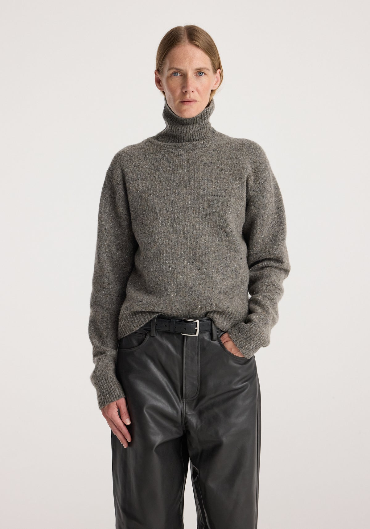 Woman wearing a grey mouliné turtleneck sweater tucked into black leather pants with a belt, standing against a plain light background, showcasing the Mouliné turtleneck | grey mouliné.