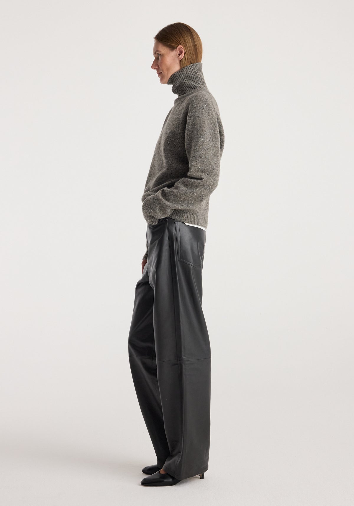Woman wearing a grey mouliné turtleneck sweater, styled with loose black leather pants and black shoes, standing in profile against a plain background, showcasing the Mouliné turtleneck | grey mouliné.