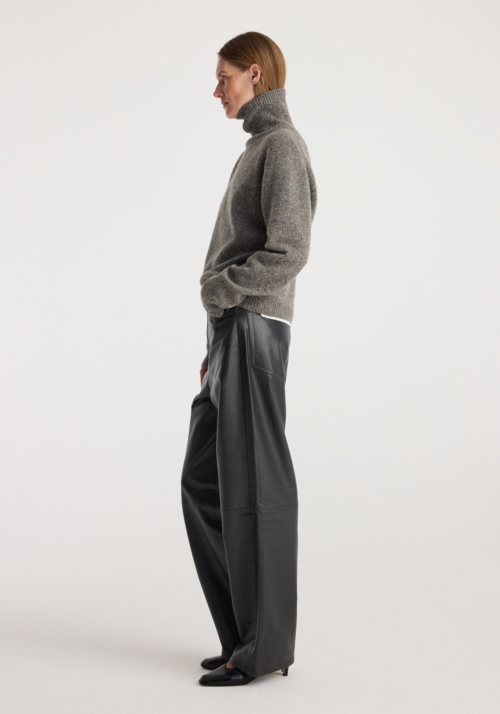 Woman wearing a grey mouliné turtleneck sweater, styled with loose black leather pants and black shoes, standing in profile against a plain background, showcasing the Mouliné turtleneck | grey mouliné.