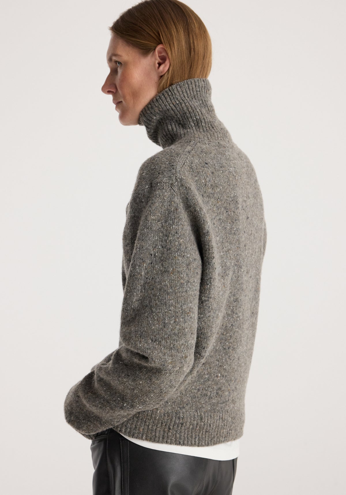 Woman wearing a grey speckled knitted high-neck sweater, styled with black pants, shown in profile against a plain light background, featuring the Mouliné turtleneck | grey mouliné.