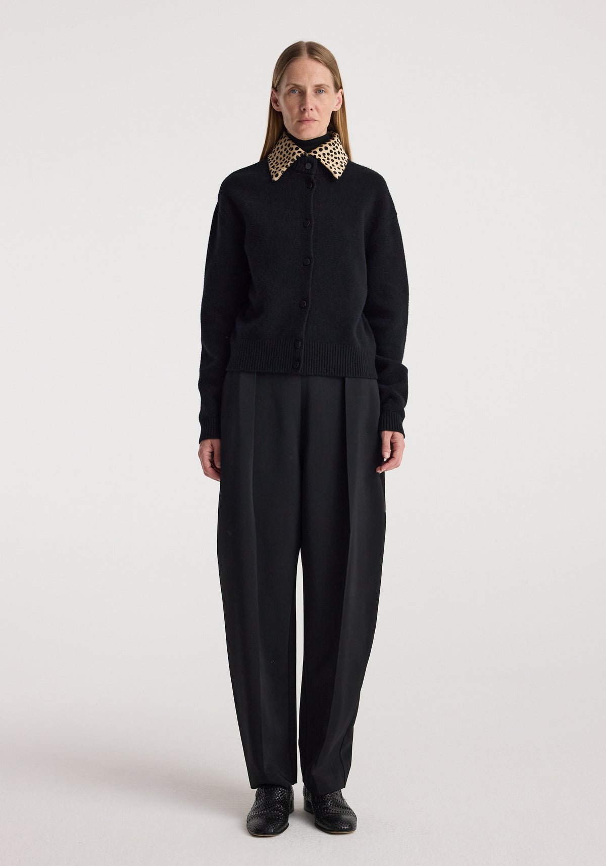 Woman wearing a black button-up cardigan with a cheetah print collar, paired with loose black trousers and black shoes, standing against a plain white background, featuring the Cheetah collar cardigan | black.