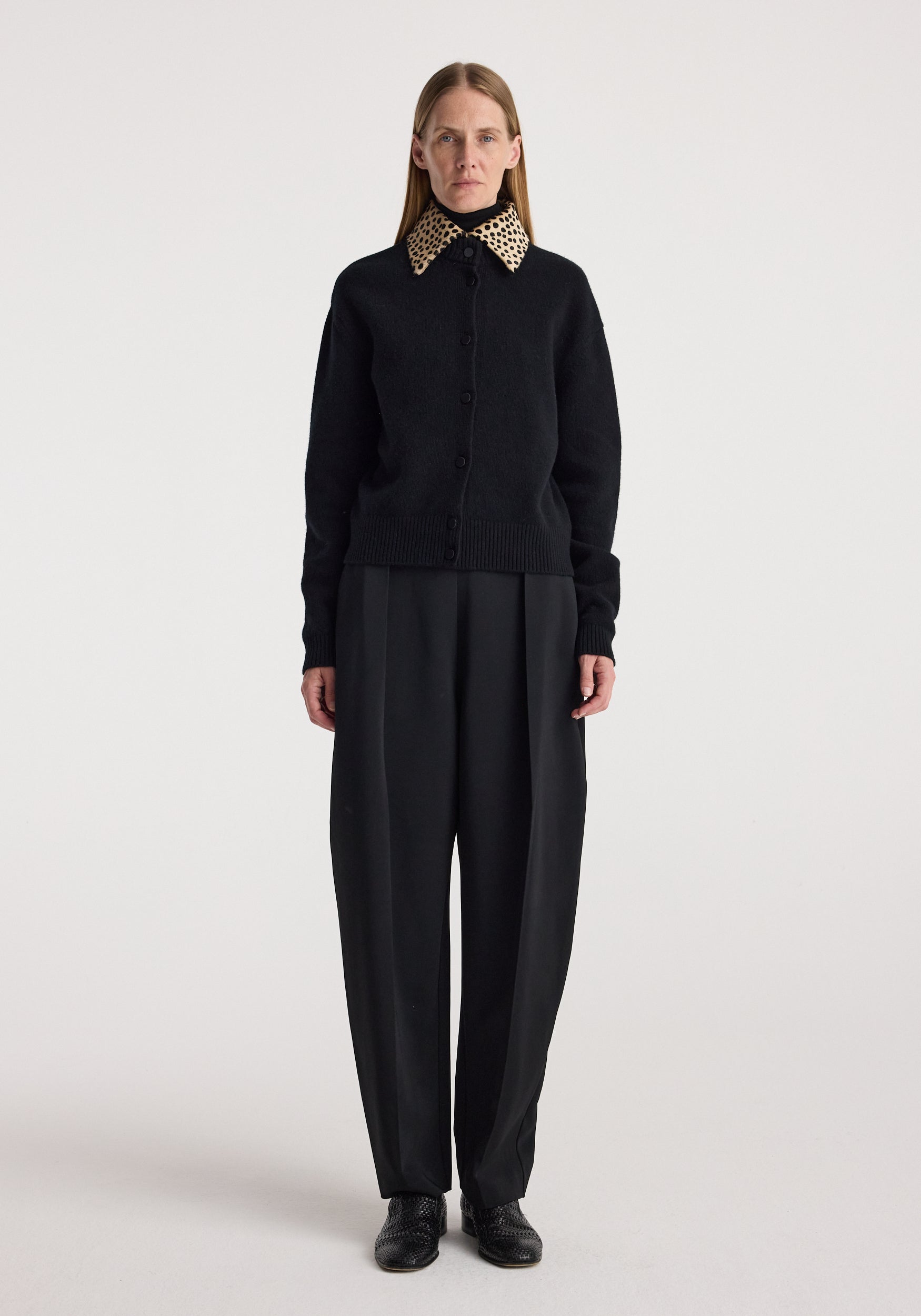 Woman wearing a black button-up cardigan with a cheetah print collar, paired with loose black trousers and black shoes, standing against a plain white background, featuring the Cheetah collar cardigan | black.