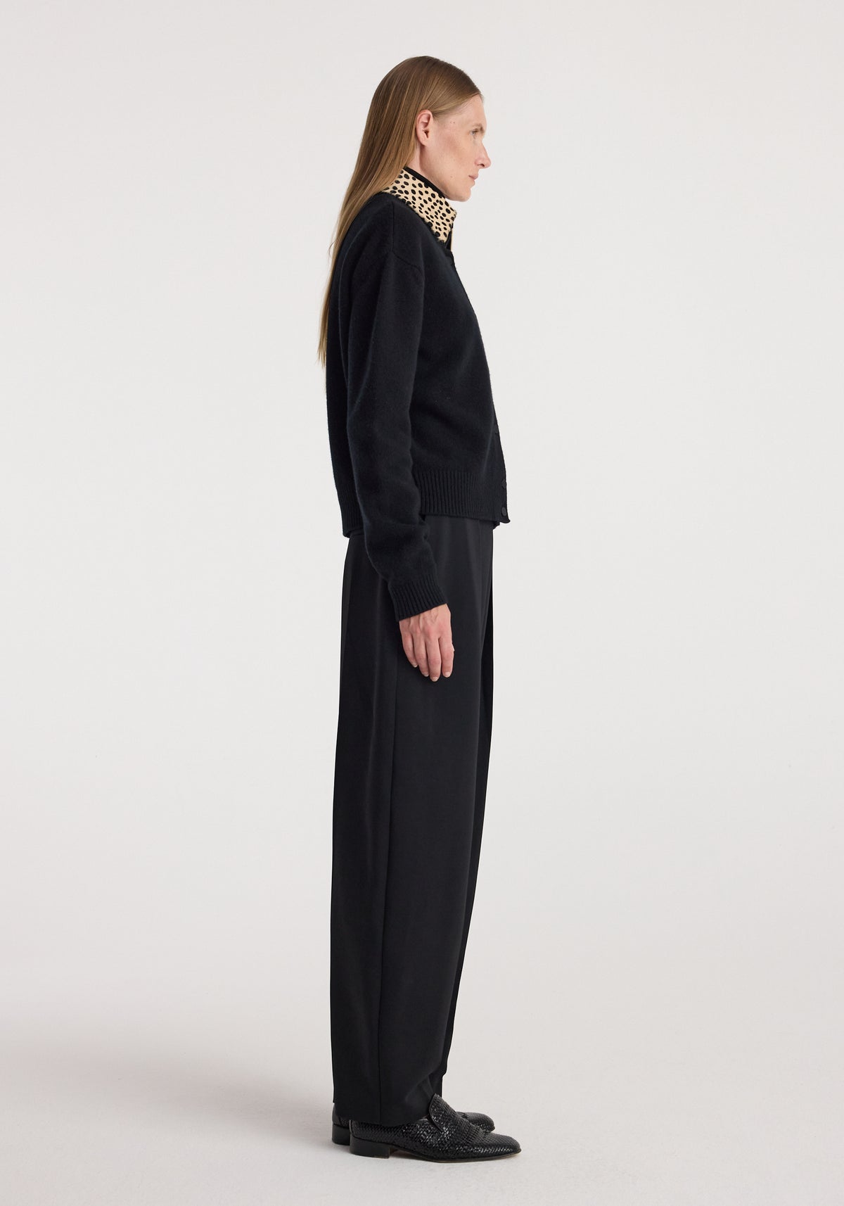 Side view of a woman wearing a black Cheetah collar cardigan | black with a cheetah print collar, paired with black wide-leg pants and black shoes against a plain background.