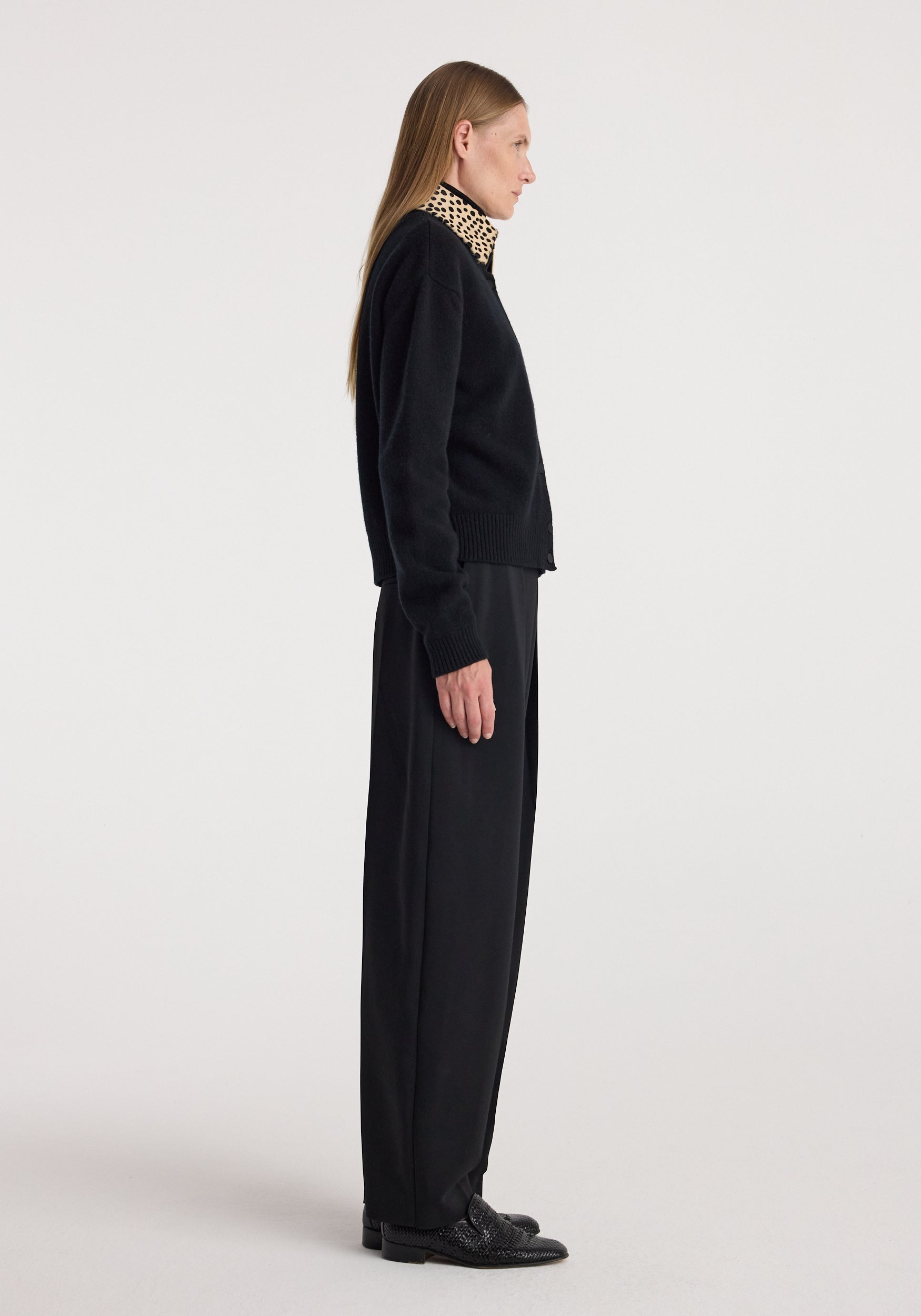 Side view of a woman wearing a black Cheetah collar cardigan | black with a cheetah print collar, paired with black wide-leg pants and black shoes against a plain background.
