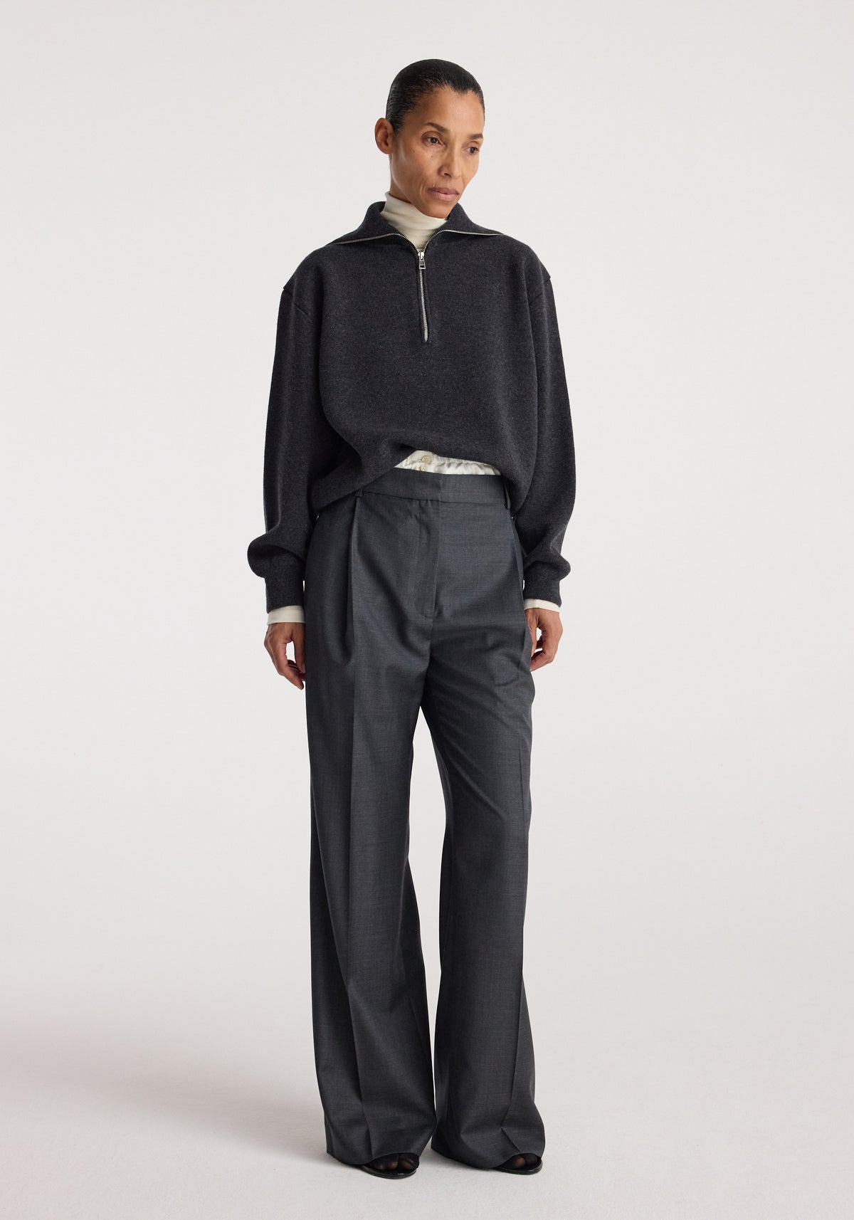 A woman wearing a charcoal Two-way zipped turtleneck layered over a light turtleneck, paired with dark wide-leg trousers, standing against a plain light background.