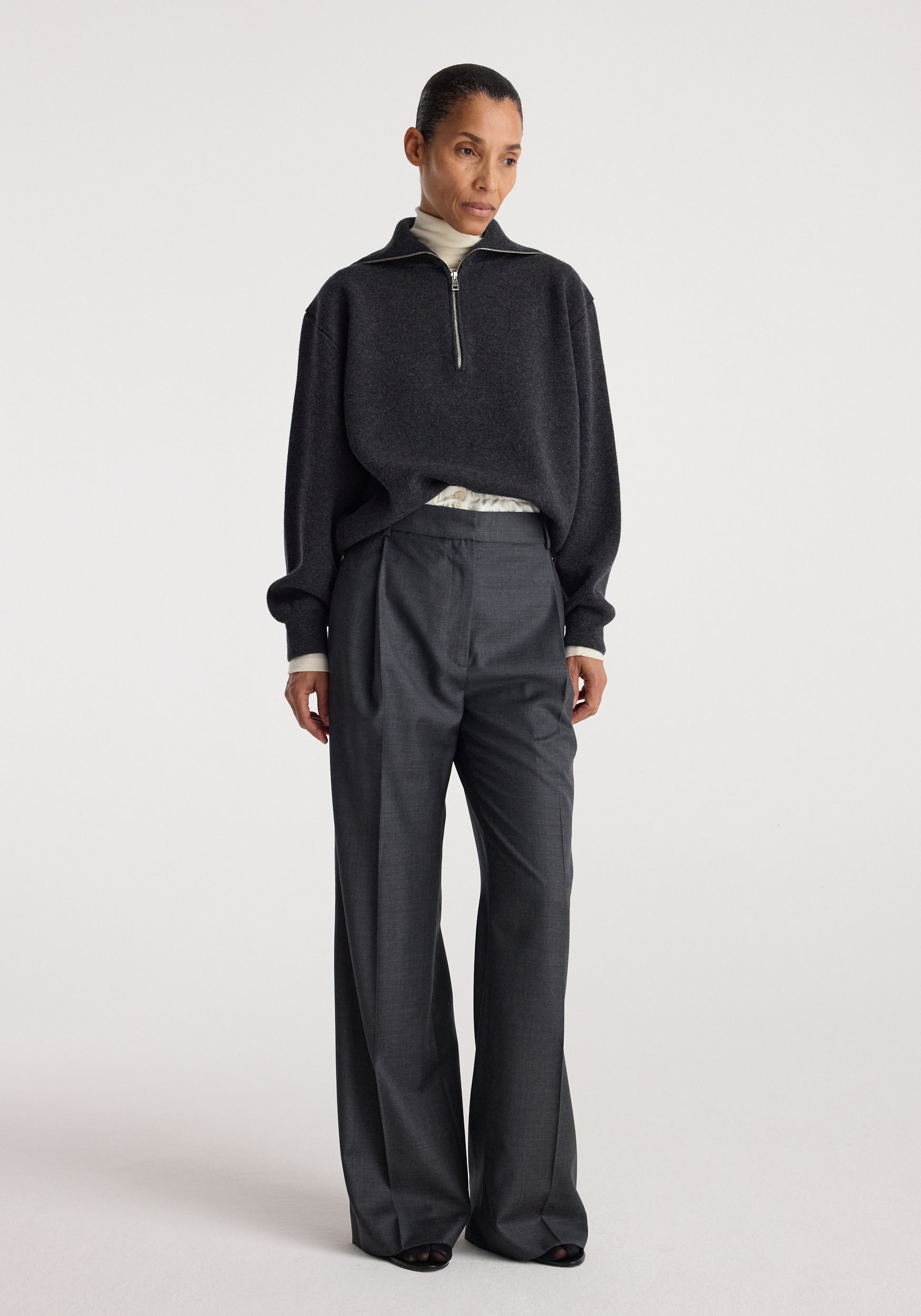 A woman wearing a charcoal Two-way zipped turtleneck layered over a light turtleneck, paired with dark wide-leg trousers, standing against a plain light background.